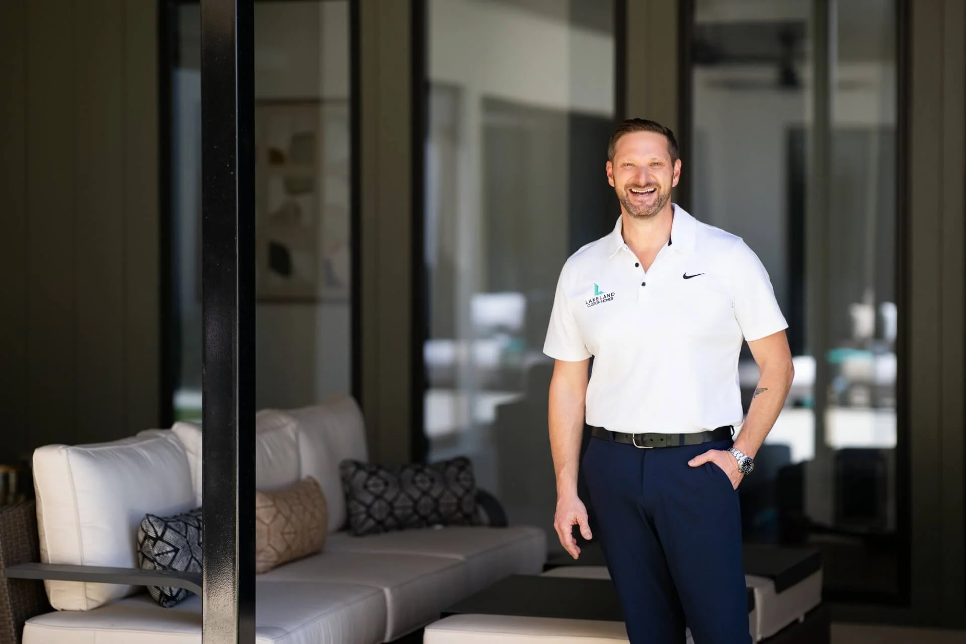 Nick Zuckerbrow with Lakeland Custom Homes in Burelson, TX with a beard and short hair smiling, dressed in a white polo shirt and navy pants, standing in a modern indoor setting with outdoor view and a white sofa with patterned pillows.