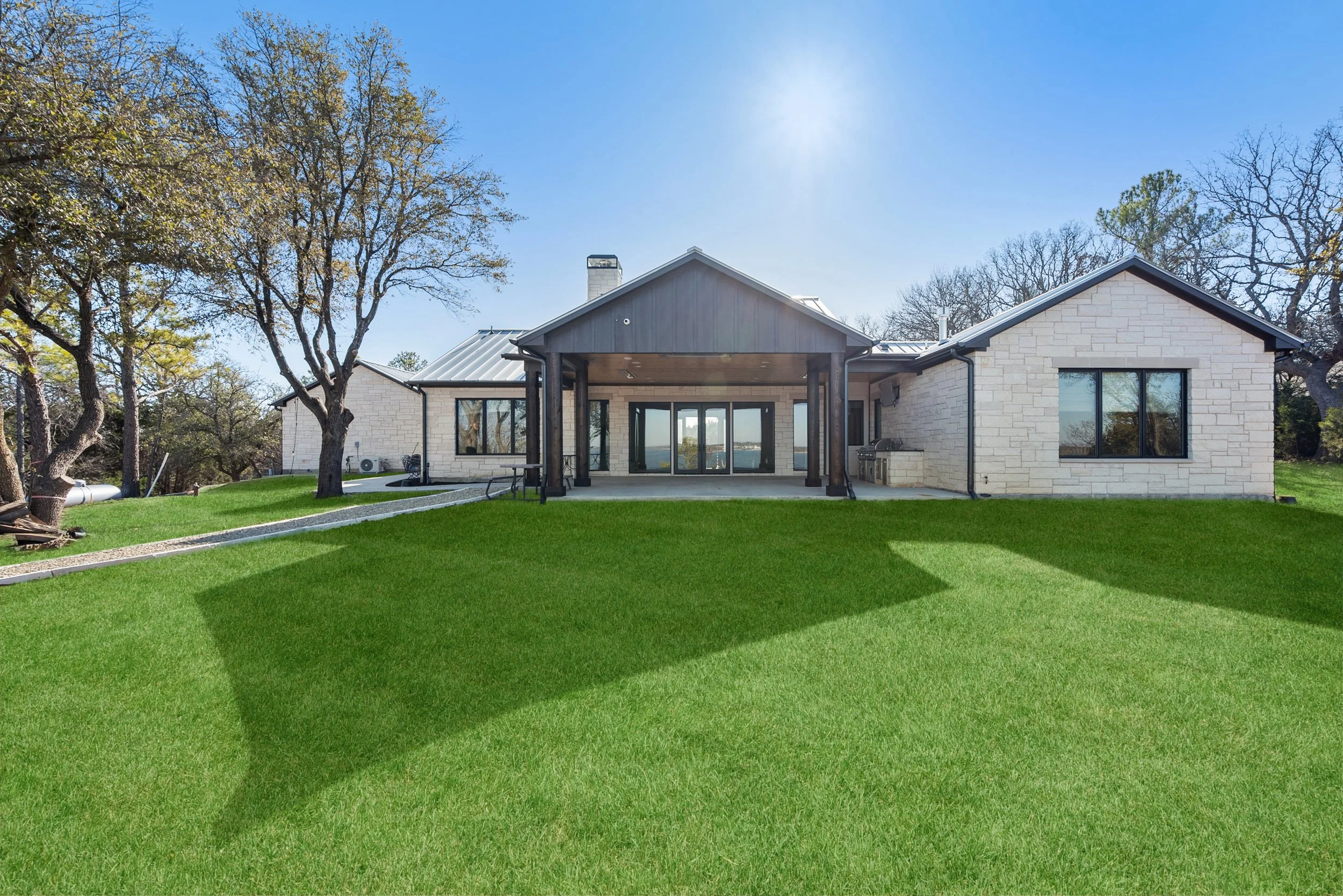 A modern single-story house with a well-maintained green lawn and trees, featuring a patio area with a grill and outdoor furniture, under a clear blue sky built by Lakeland Custom Homes.
