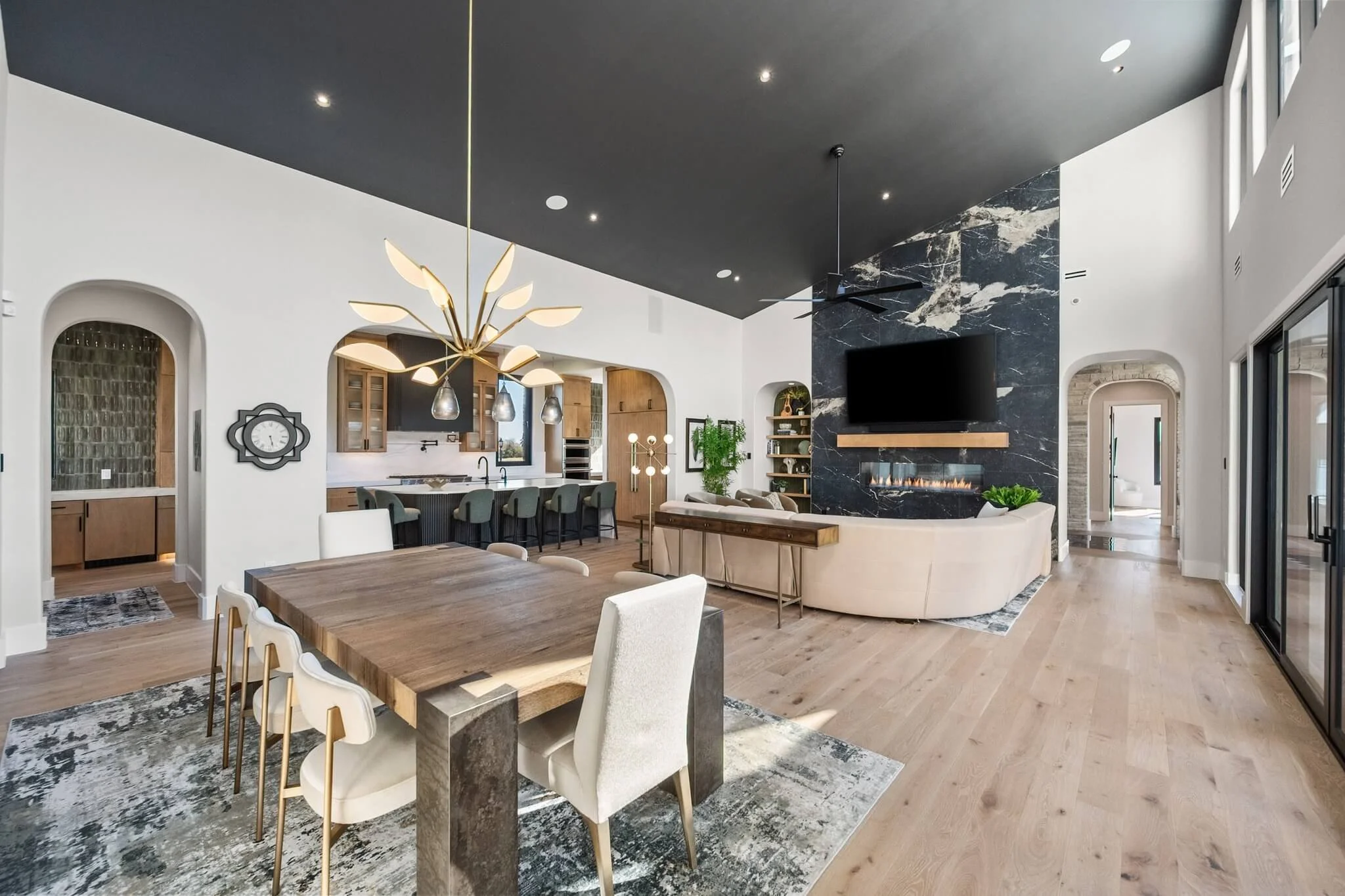 Modern open-concept living and dining area with a black ceiling, a unique chandelier, a fireplace with a black marble surround, a flat-screen TV, a wooden dining table with white chairs, and a kitchen with barstools, wooden cabinets, and large window