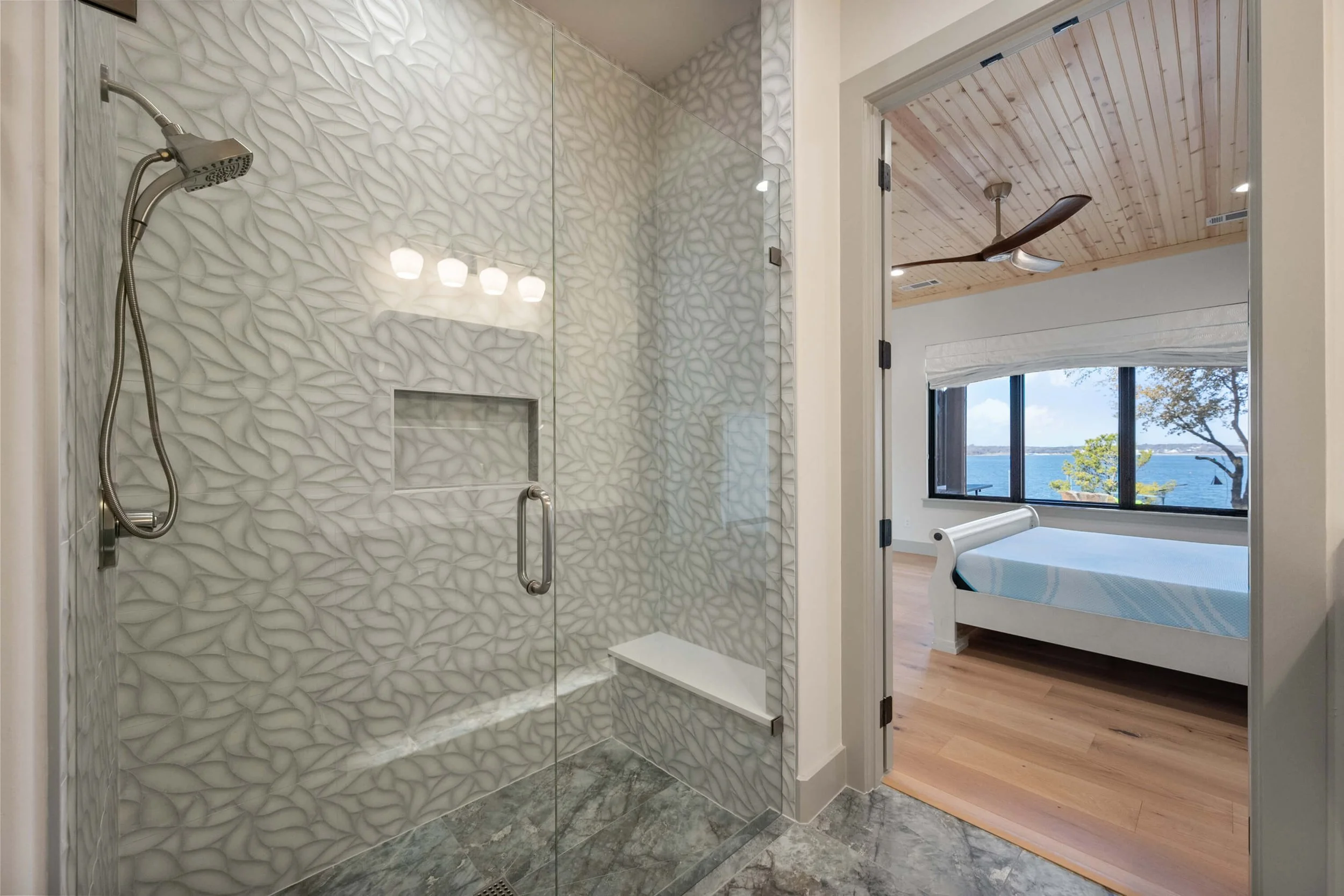 Bathroom shower with textured tile walls and glass door, view into a bedroom with a bed, large window, and lake view built by Lakeland Custom Homes.