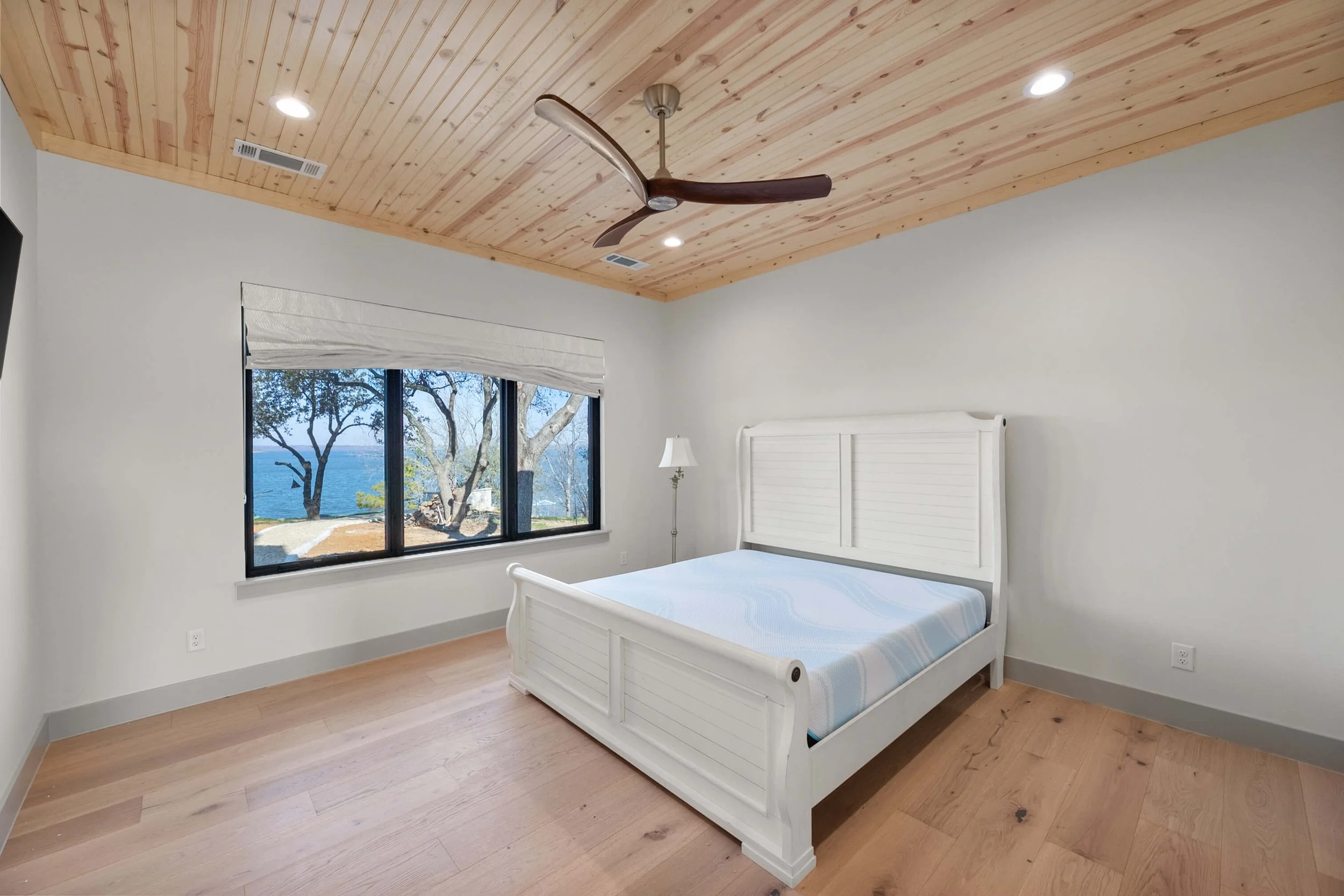 Empty bedroom with white bed, floor lamp, large window with view of trees and water, wooden ceiling with ceiling fan, hardwood floor.