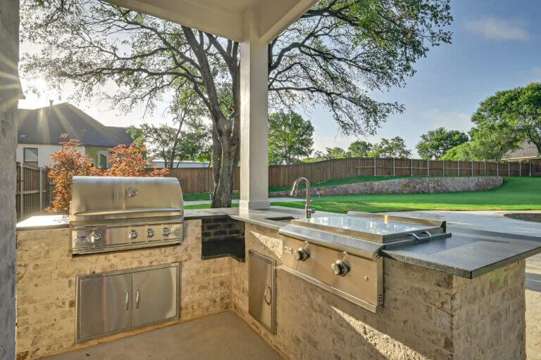 Outdoor kitchen with a built-in grill, sink, and countertop, set on a stone base, under a covered patio with a view of a backyard with trees and a fence.