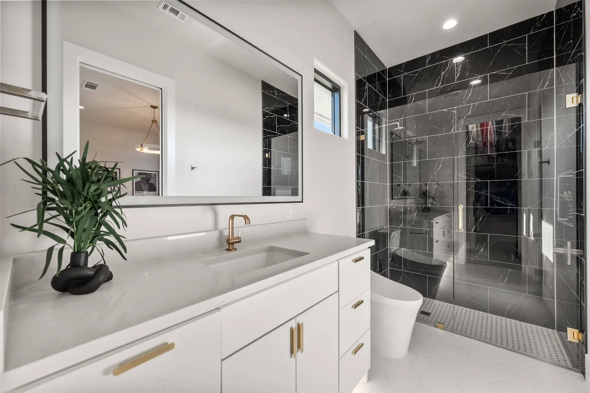 Modern bathroom with white vanity, gold hardware, large mirror, black marble shower with gold fixtures, small windows, and white tile floor.