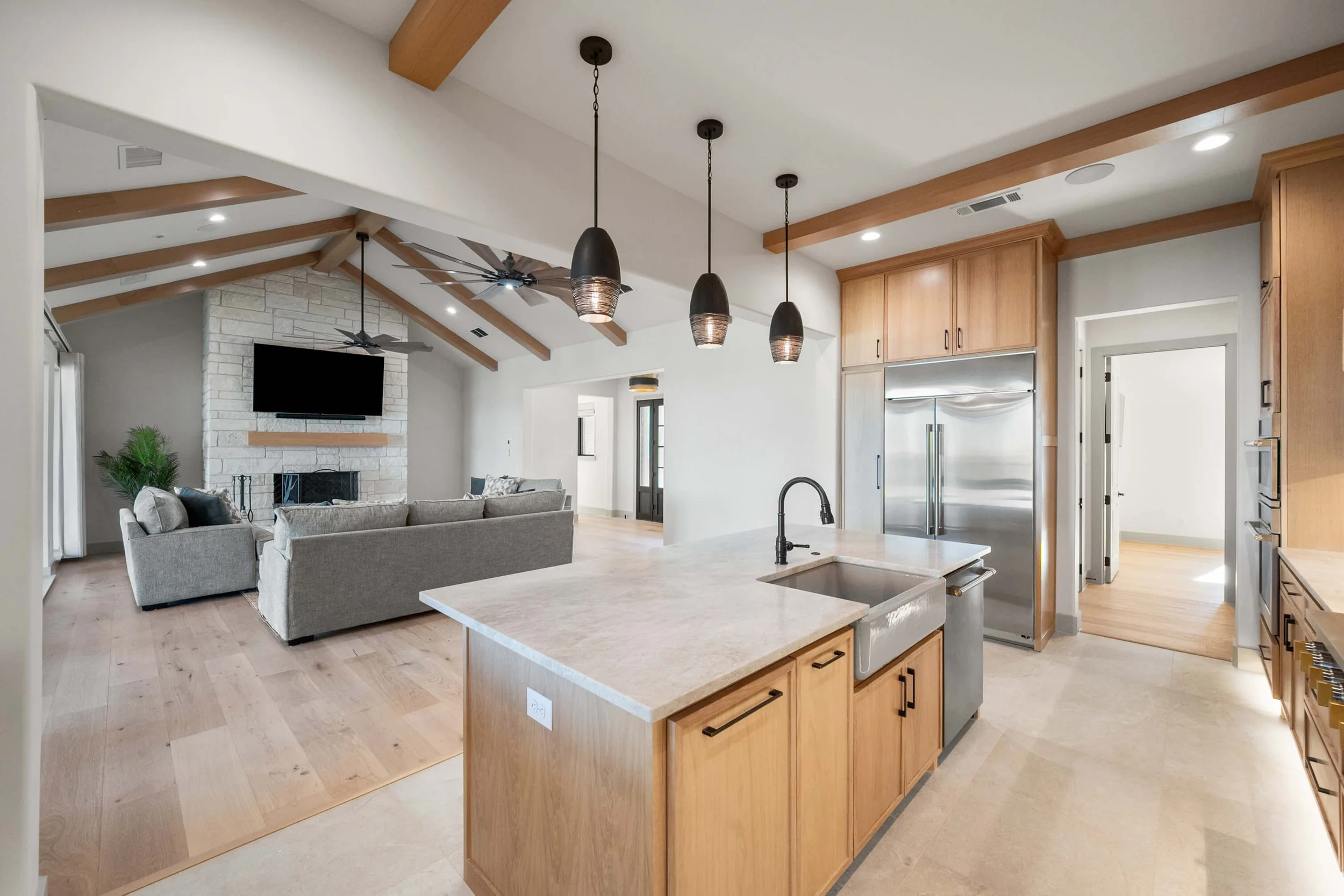 Open-concept kitchen and living room with vaulted ceiling, wooden beams, and a stone fireplace. Kitchen features a central island, stainless steel refrigerator, and pendant lighting.