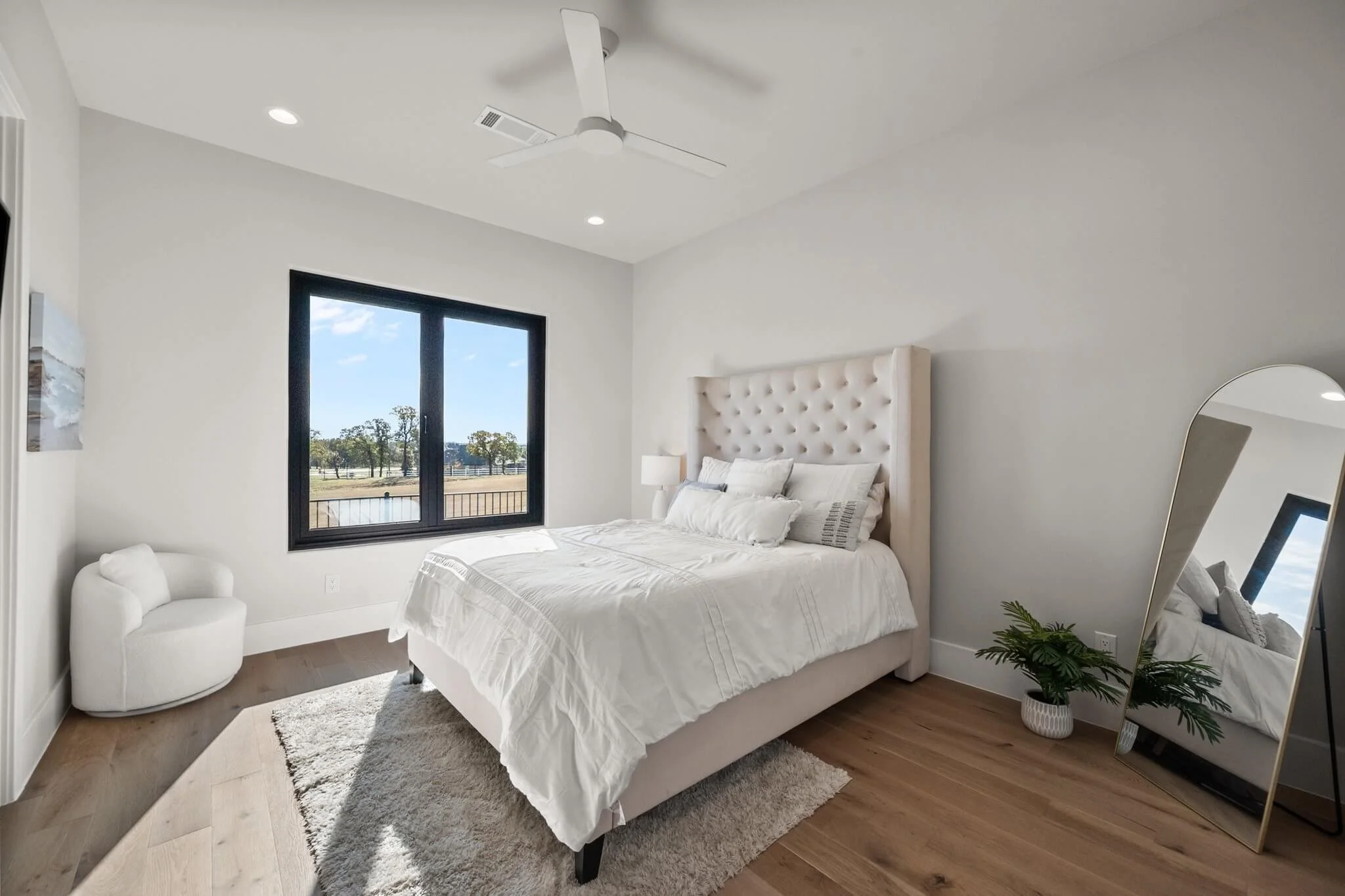 A modern bedroom with a large bed with white bedding and a tufted beige headboard, a small white chair in corner, a potted plant, a curved full-length mirror, and a window showing trees outside, all with minimalist decor.