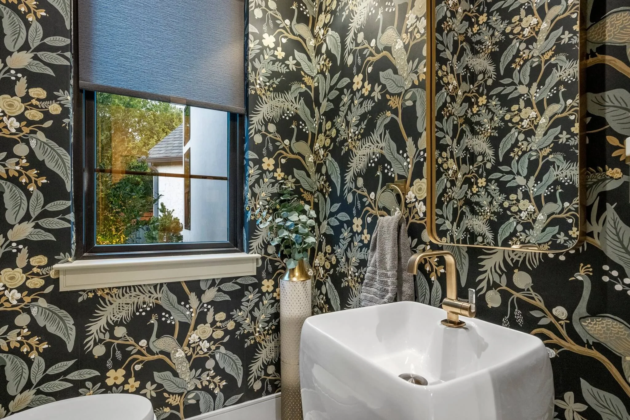 A bathroom with black and gold floral wallpaper, a window with a gray shade showing trees outside, a white rectangular sink with a gold faucet, a gold vase with greenery, and a miniature mirror  built by Lakeland Custom Homes..