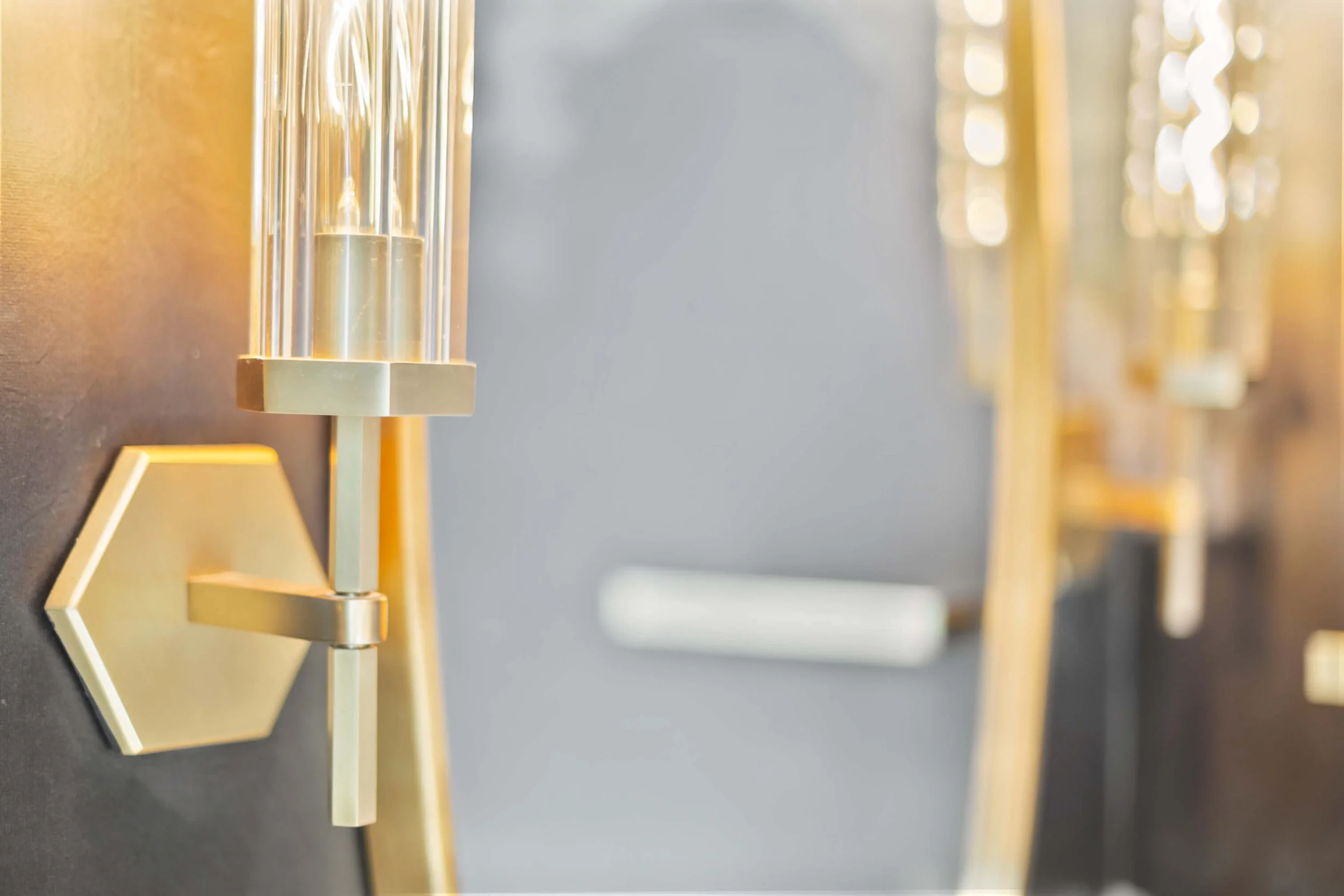 Close-up of a modern wall sconce with a gold finish and clear glass tubes, mounted on a dark wall with a blurred background.
