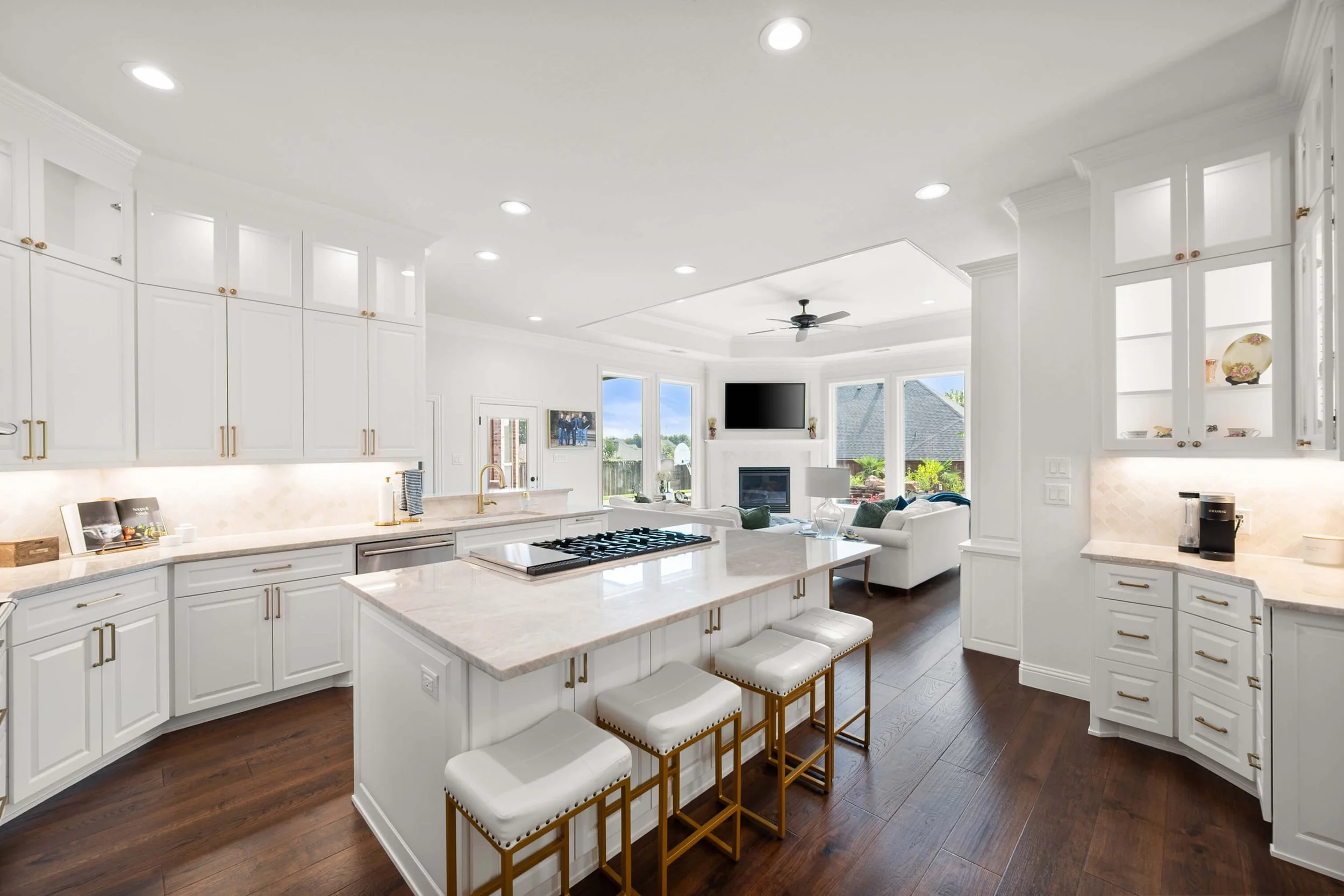 Bright, white kitchen with marble countertops, white cabinets, and gold hardware, open to a living room with large windows, a ceiling fan, and a fireplace.