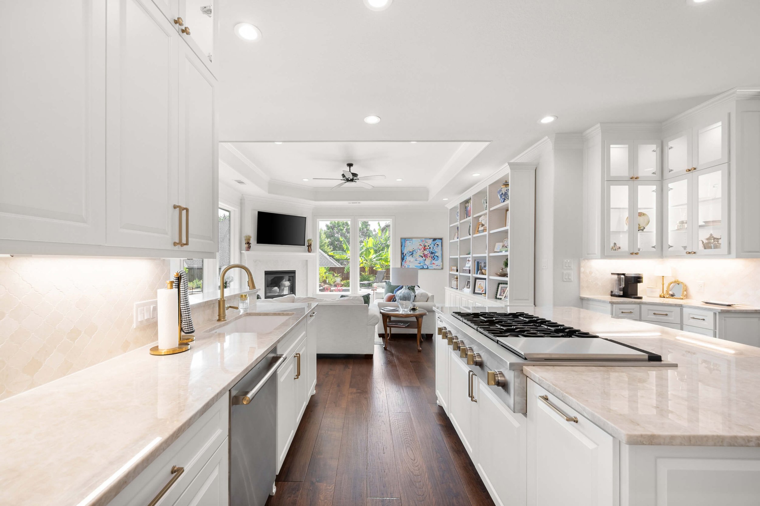 Bright open-concept kitchen with white cabinets, marble countertops, a gas stove, and a view into the living room with a fireplace, TV, and large windows.