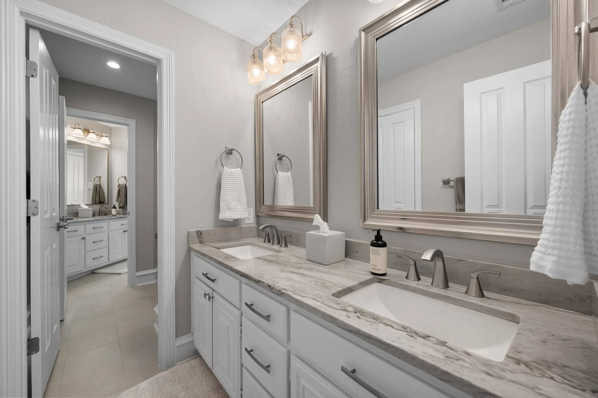 Bathroom with double vanity sinks, large mirrors, and wall-mounted light fixtures, with a doorway leading to a separate area with additional bathroom features.