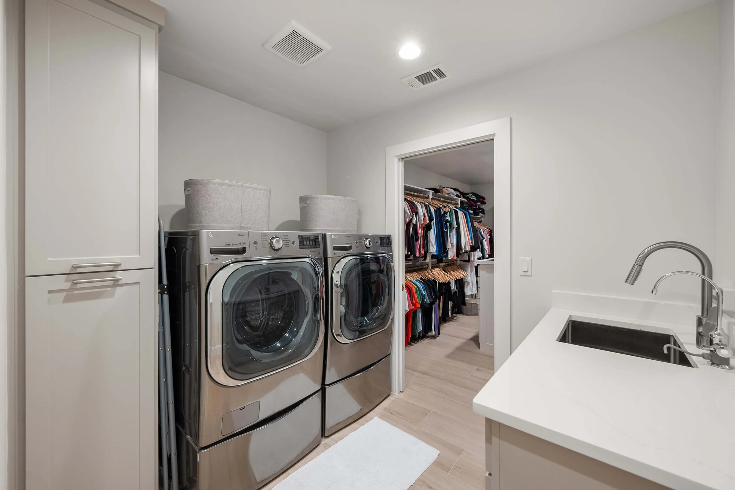 Laundry room with matching front-loading washer and dryer, storage cabinet, laundry baskets, part of a sink with a faucet, and an adjacent walk-in closet with clothes on hangers and shelves.