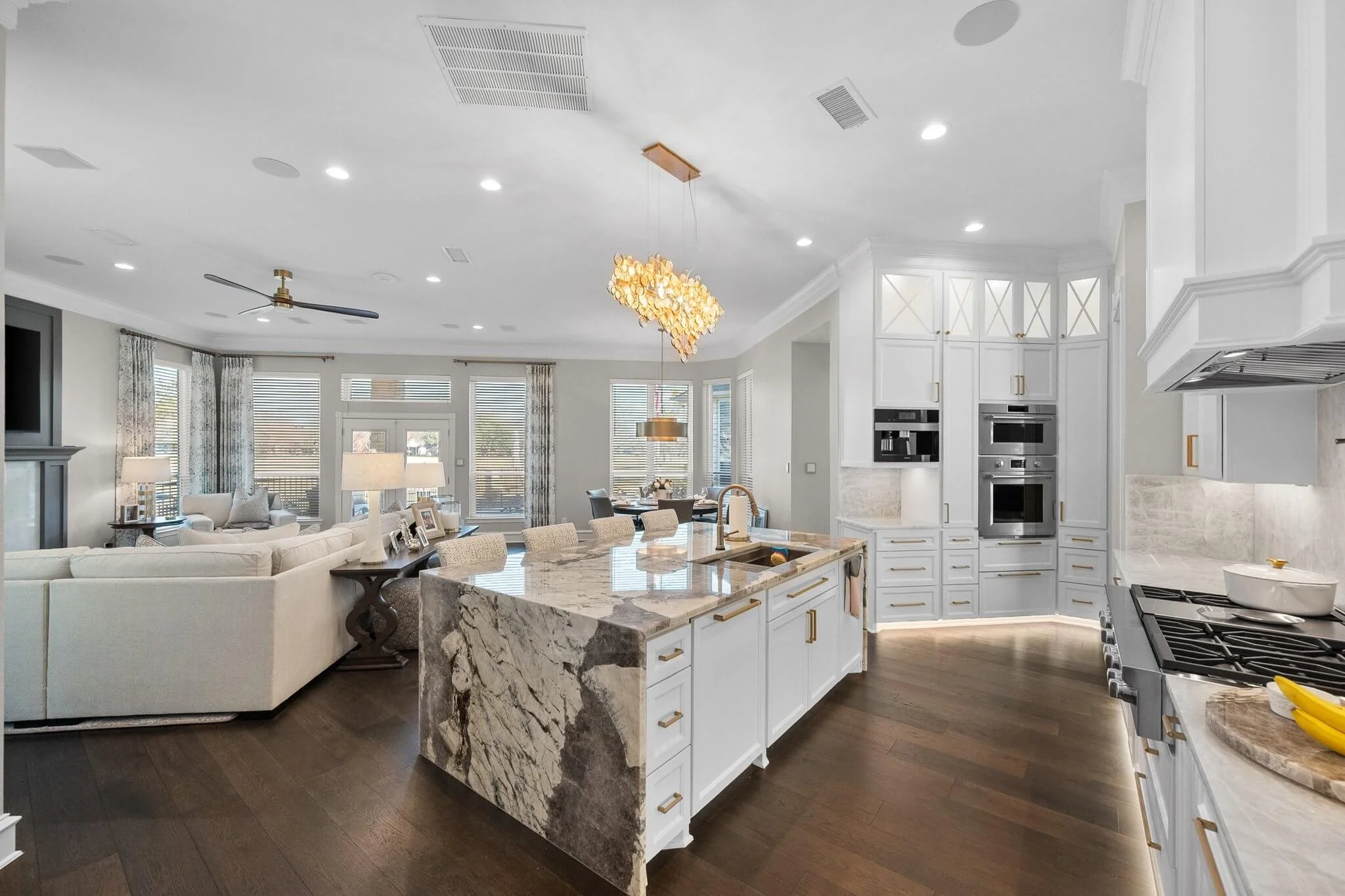 Open-concept kitchen and living room with white cabinetry, marble island, dark hardwood floors, and large windows with curtains.