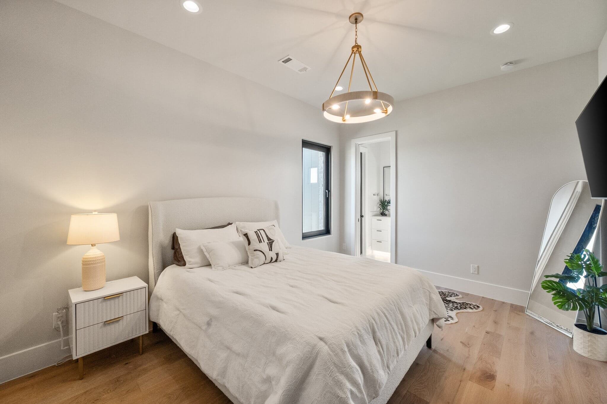 Bedroom with white walls, a white bed with a beige headboard, a white nightstand with a beige lamp, a window, a doorway leading to a bathroom with a plant, a large mirror leaning against the wall, a wall-mounted TV, and a potted plant in the corner. 