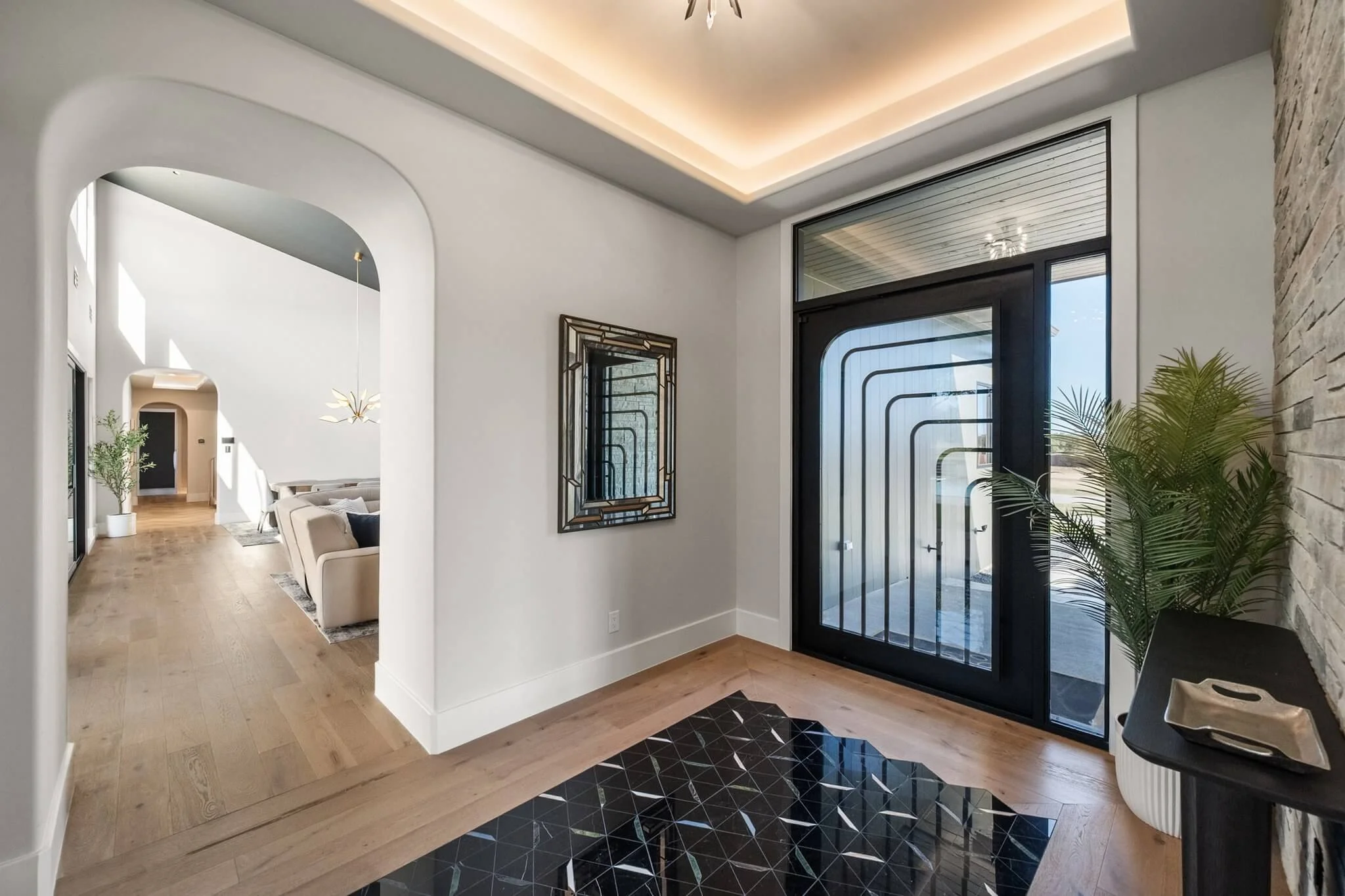 Entryway with black door featuring geometric window design, wooden flooring, and a potted plant on a black side table. Past the curved archway, a living room with beige sofa, wall mirror, and hanging light fixture are visible.