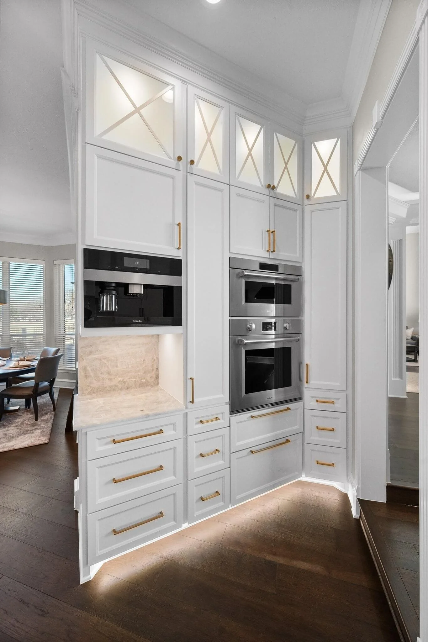 White built-in kitchen cabinets with gold handles, a built-in coffee maker, and double wall ovens, in a modern kitchen with dark hardwood floors.