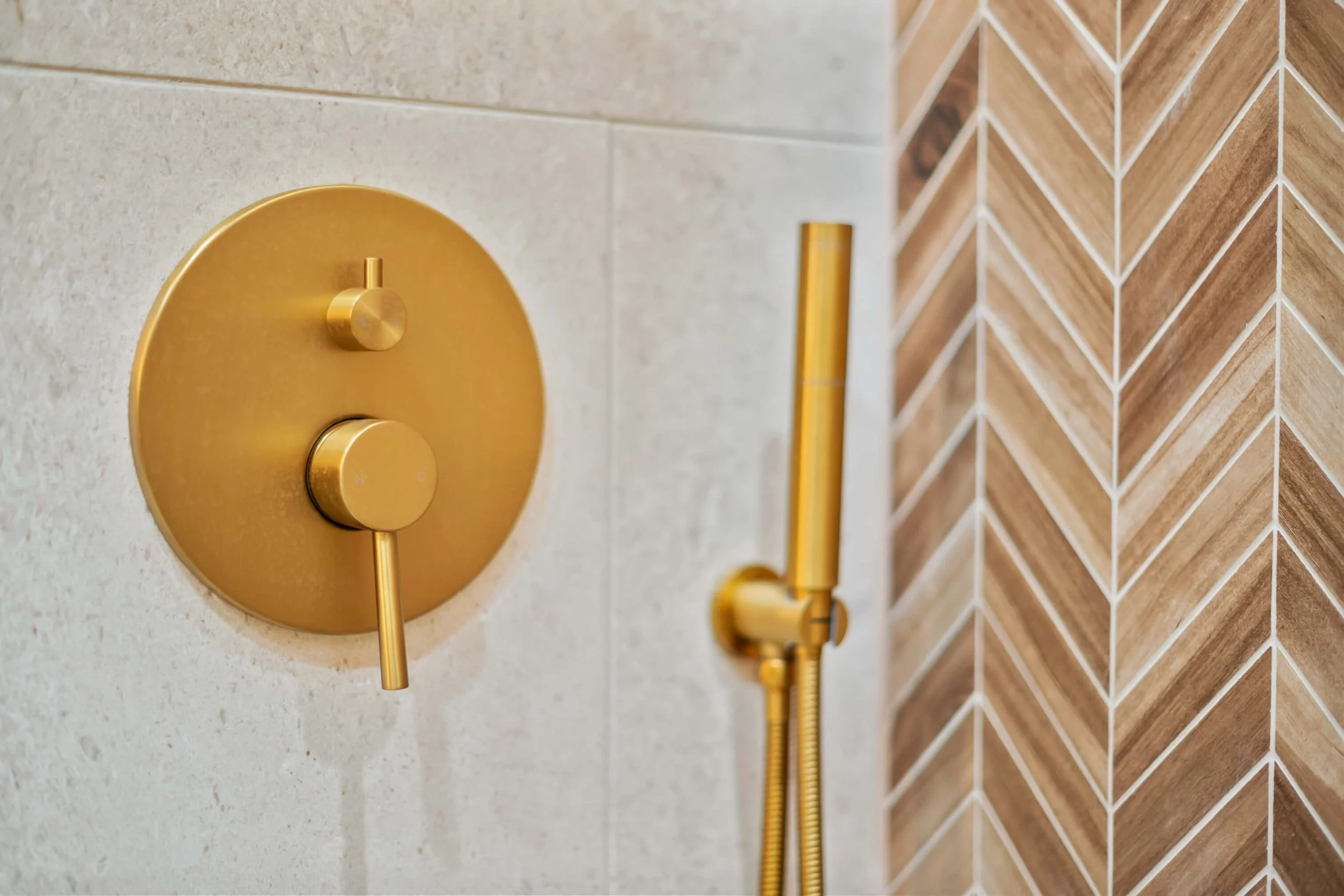 Close-up of a modern shower wall with a gold shower control panel and a handheld showerhead against beige and wood-patterned tiles built by Lakeland Custom Homes..