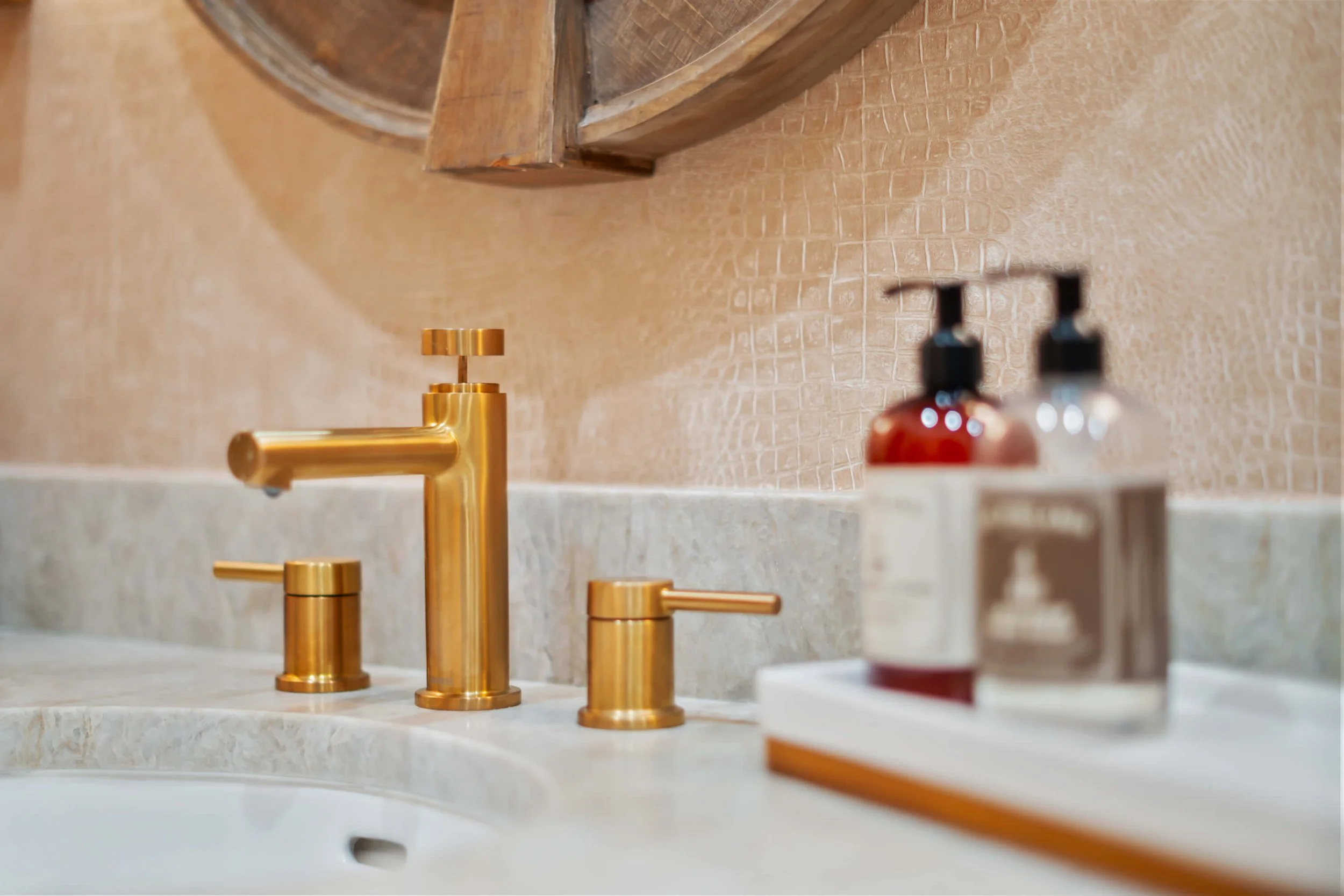 Close-up of a gold bathroom faucet with two gold handles on a beige marble sink, with soap dispensers in the foreground and a textured beige wall with a round mirror above built by Lakeland Custom Homes..