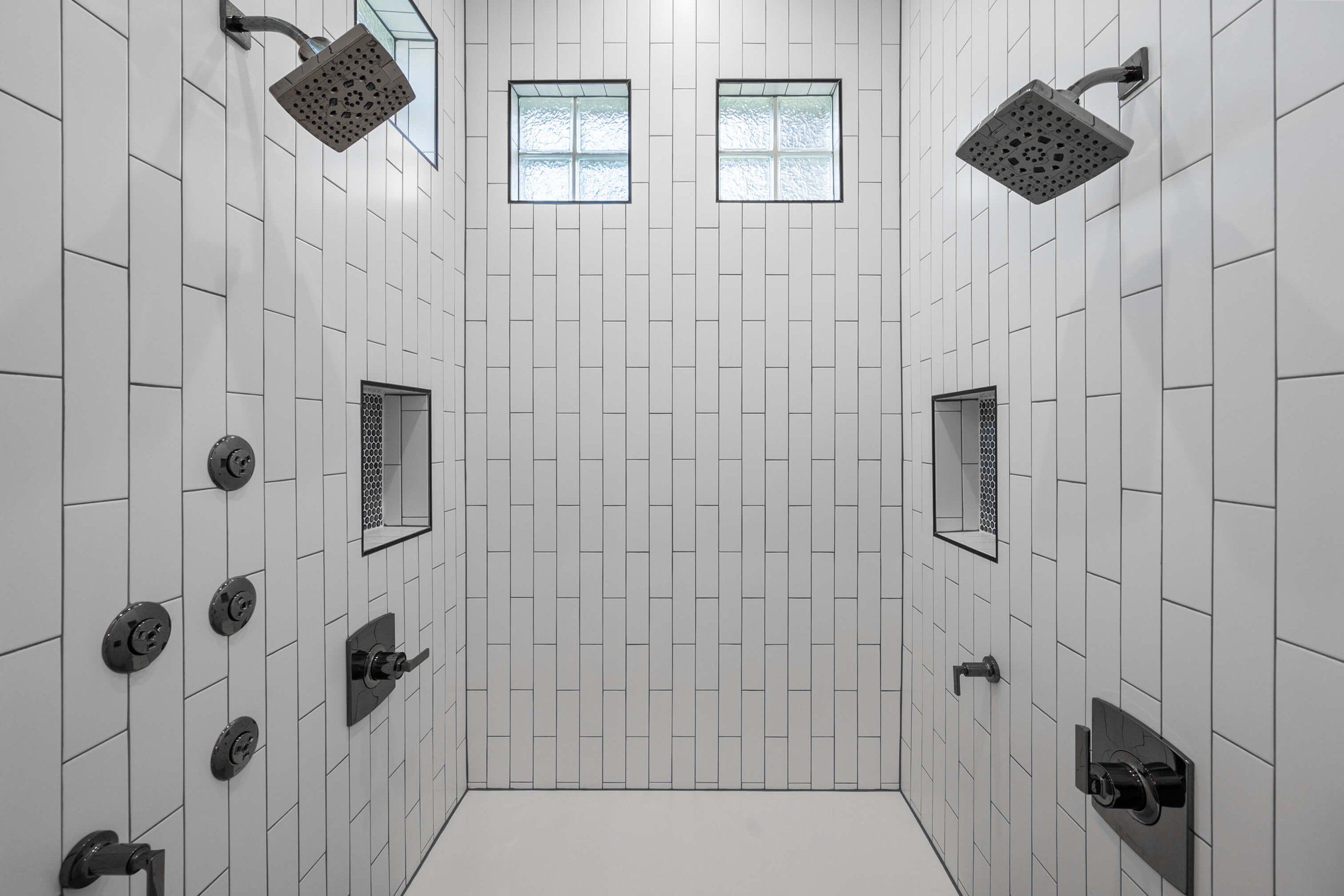 A modern shower with white subway tile walls, two showerheads on opposite sides, built-in wall niches, and two small windows at the top letting in natural light.
