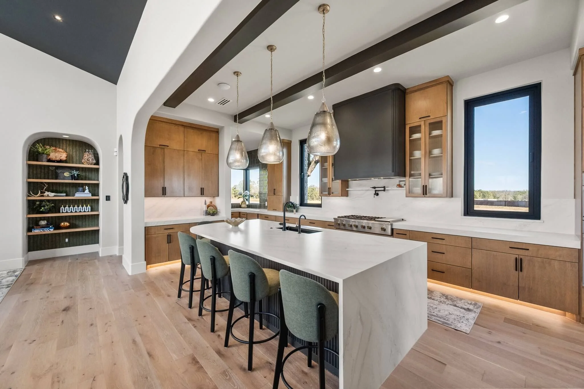 Modern kitchen with wooden cabinets, white countertops, black window frames, and a large island with four green upholstered chairs and pendant lights.