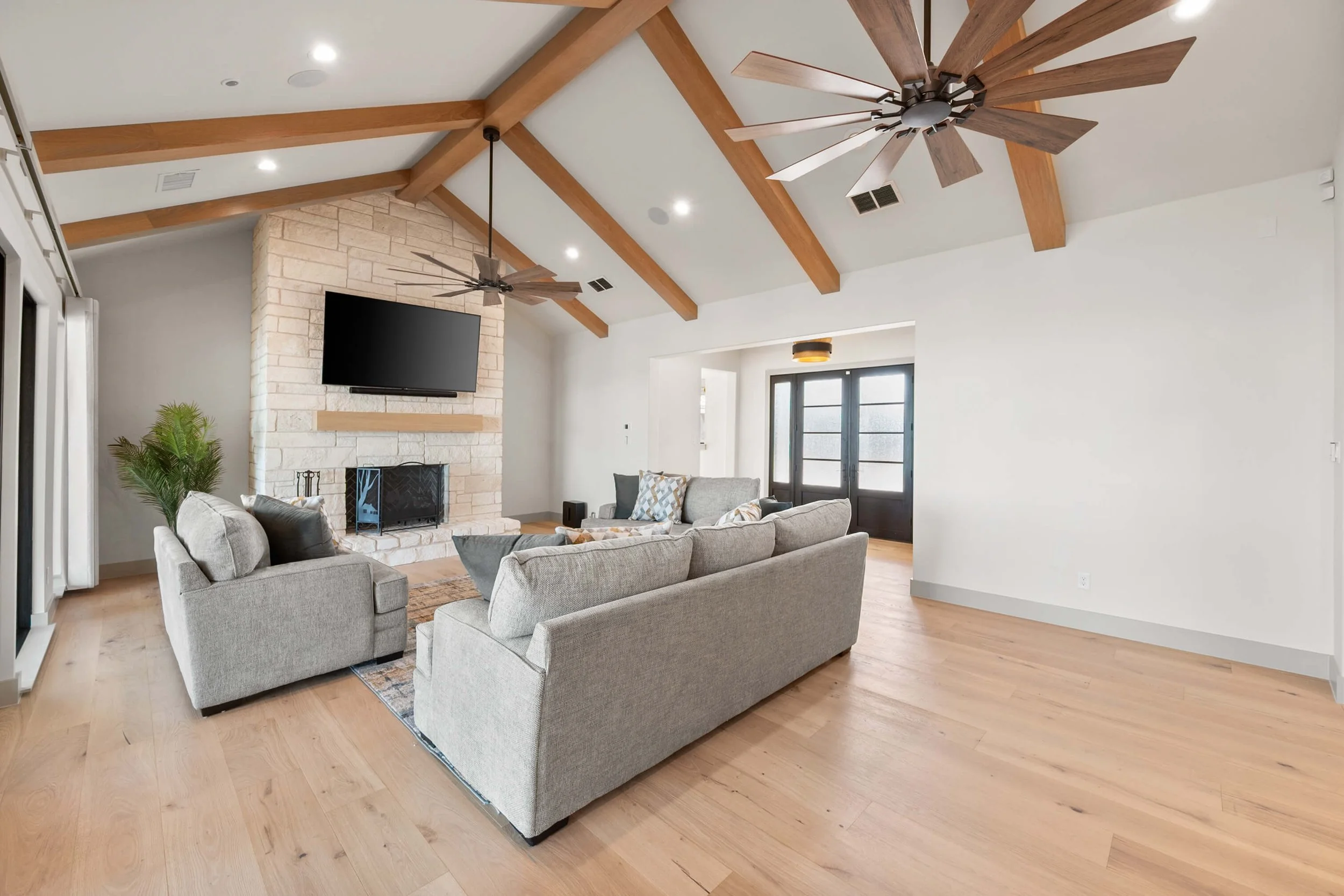 Living room with a stone fireplace, a wall-mounted TV, wooden ceiling beams, two ceiling fans, a large gray sectional sofa, a smaller matching armchair, a glass coffee table, light-colored hardwood floors, and black front door with glass panels