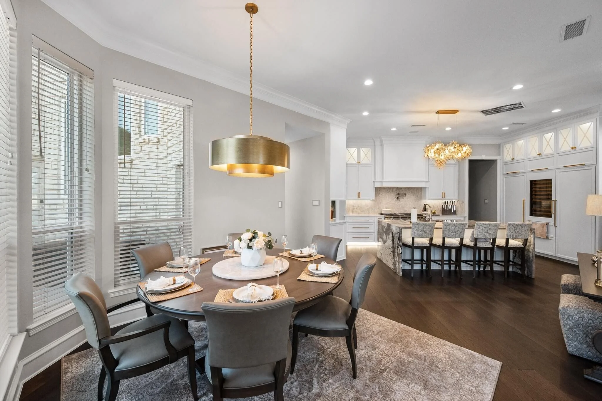 Modern dining area with a round table set for six, featuring white plates, napkins, and wine glasses, with a gold pendant light hanging above. The space is adjacent to a white kitchen with marble countertops and bar stools, illuminated by elegant lig