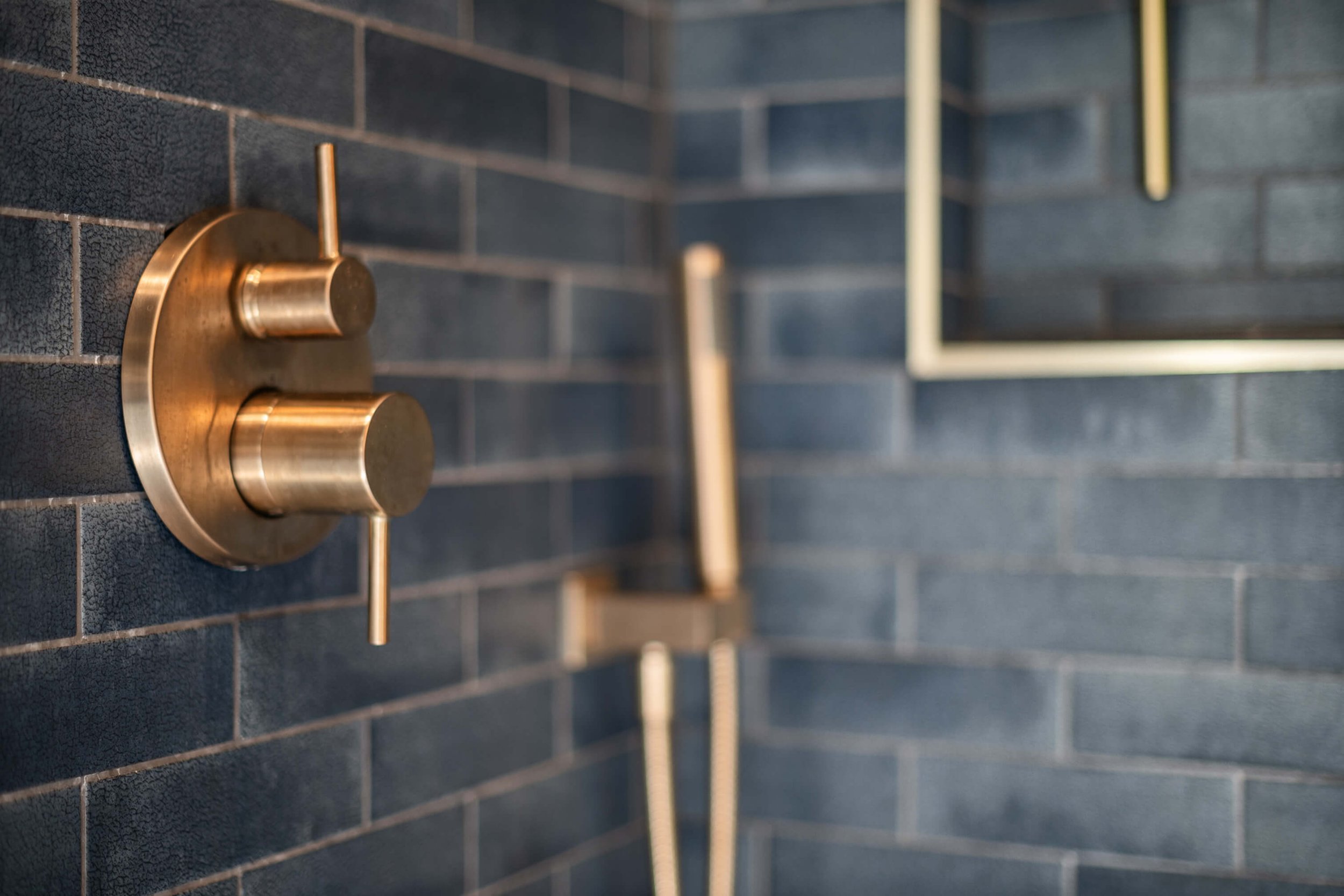 Close-up of gold-colored shower control handle mounted on a dark tiled wall in a modern bathroom built by Lakeland Custom Homes..
