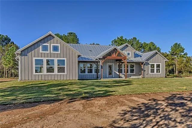 New modern house with gray siding, multiple windows, a metal roof, and a front porch surrounded by grass and trees in the background.