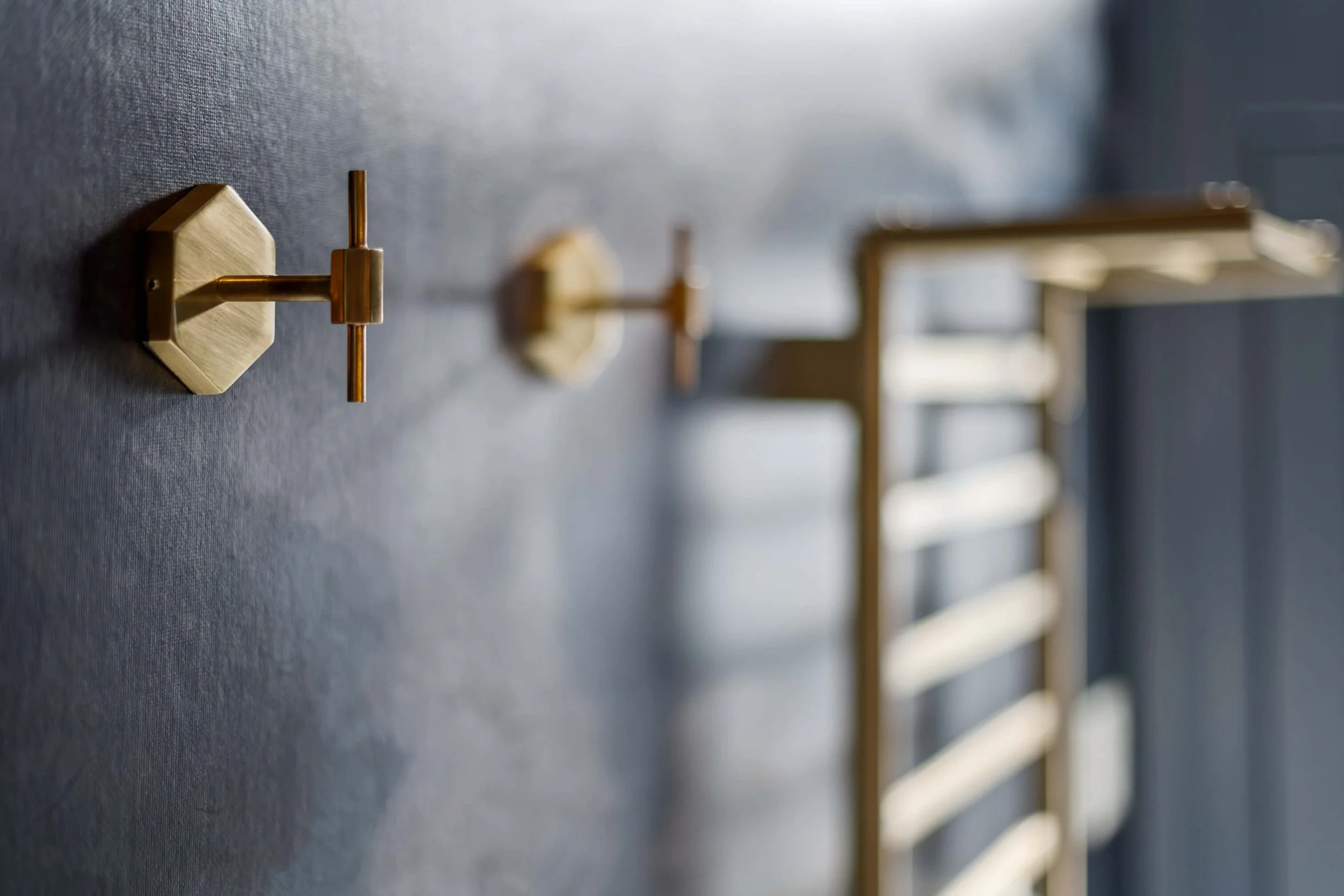 Close-up of brass wall hooks mounted on a dark gray textured wall, with a blurred wooden ladder or rack in the background.