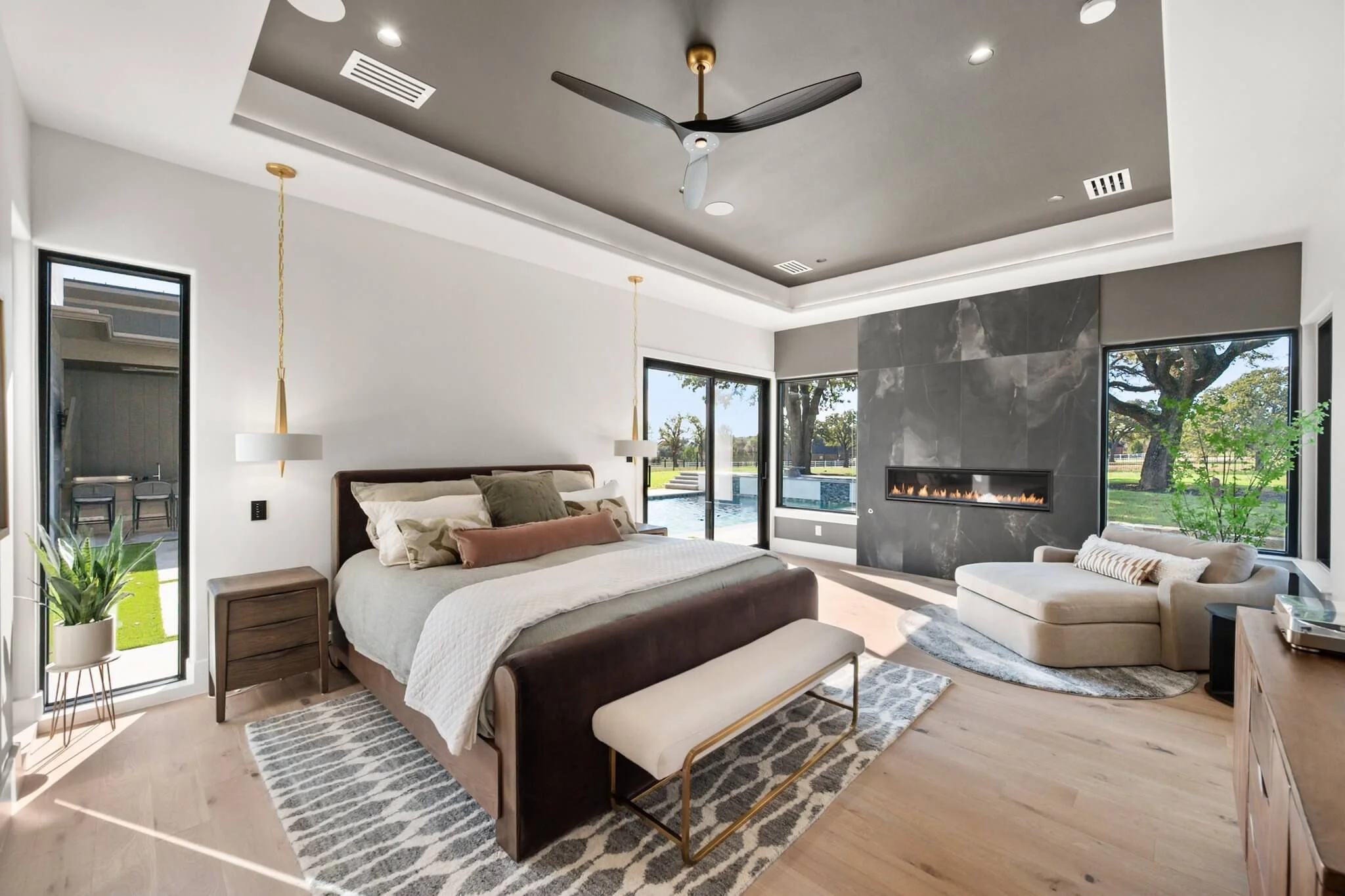 Modern bedroom with a large bed, pendant lighting, a bench at the foot of the bed, a side table, a window with sliding doors leading to an outside yard, a fireplace in the gray wall, a beige curved sofa, a round rug, a ceiling fan, and large windows 