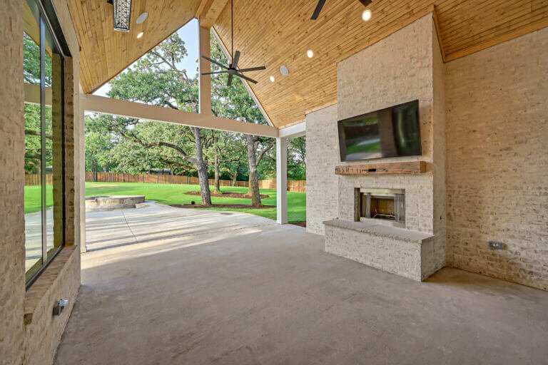 Inside view of a modern covered patio with a stone fireplace and mounted TV, overlooking a backyard with trees and a fire pit.