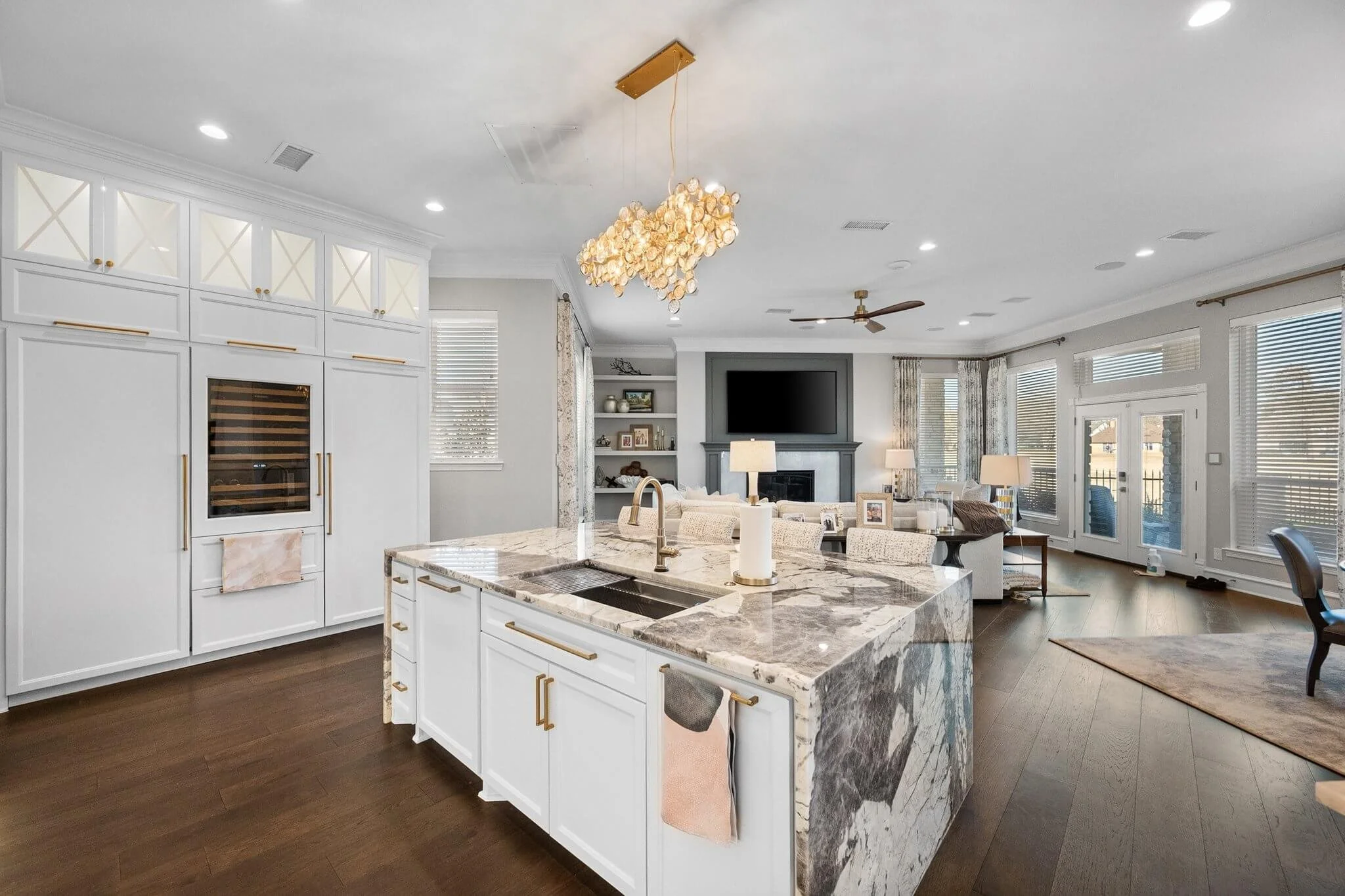 Open-concept living room and kitchen with white cabinetry, marble island, dark hardwood floors, and large windows with blinds.