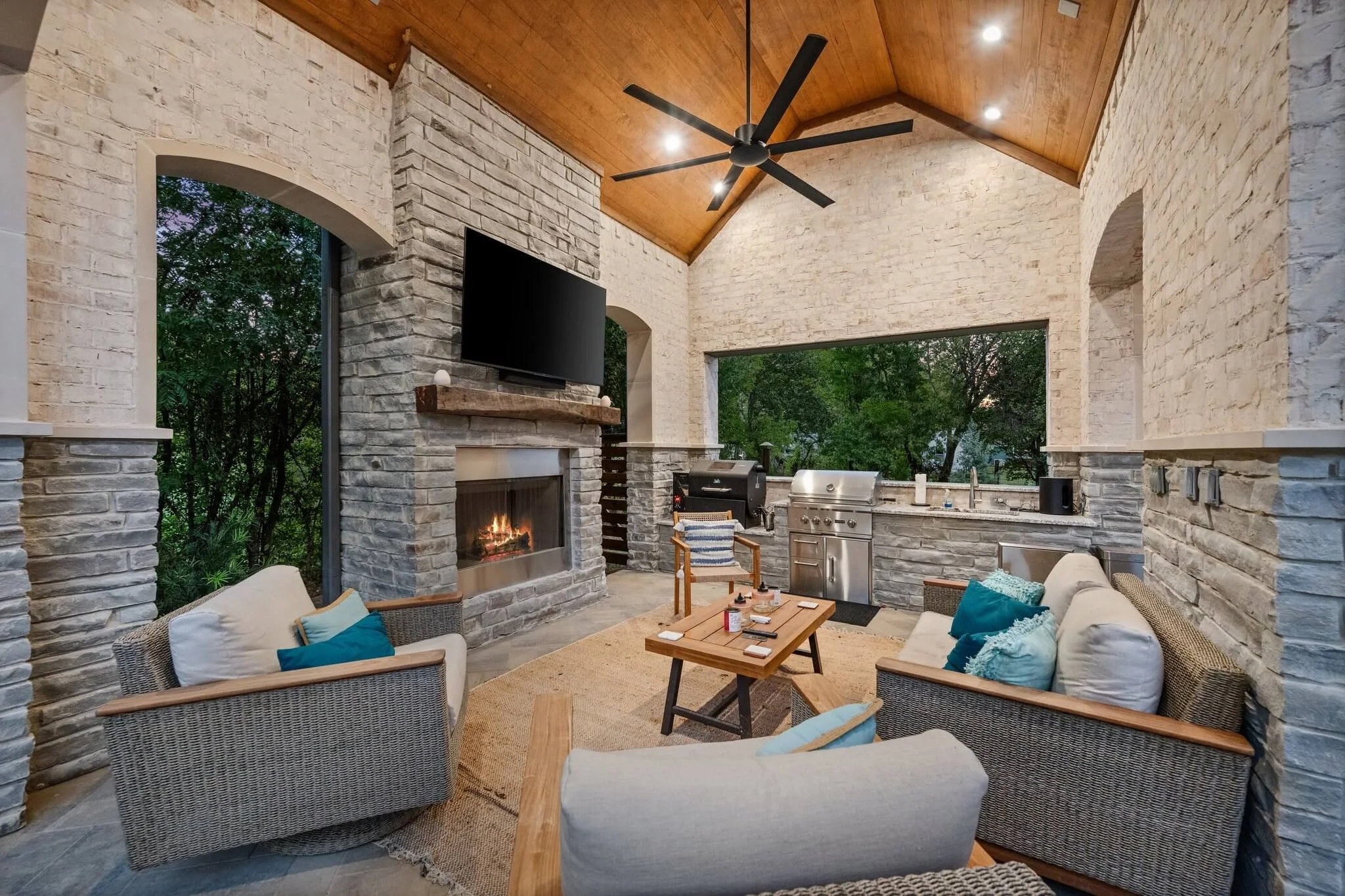 Indoor outdoor living area with a stone fireplace, a large flat-screen TV, a ceiling fan, and outdoor kitchen appliances. There are cozy sofas with cushions around a wooden coffee table, overlooking greenery outside in Colleyville, TX.