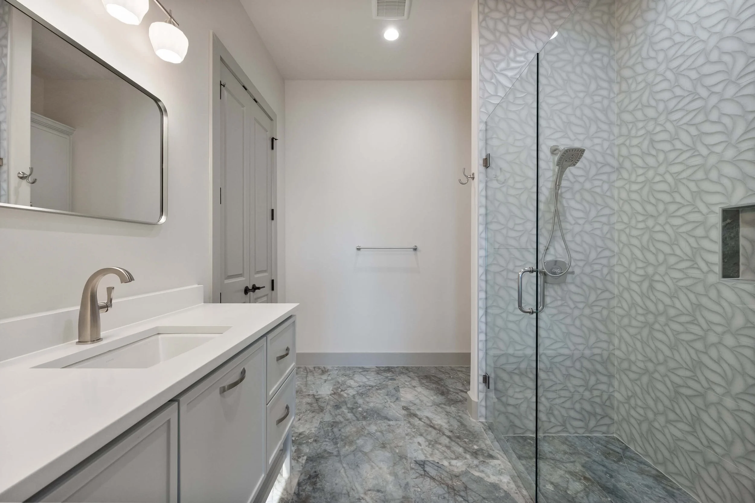 Modern bathroom with white vanity, marble floor, and glass-enclosed shower with textured white tile walls and rainfall showerhead.