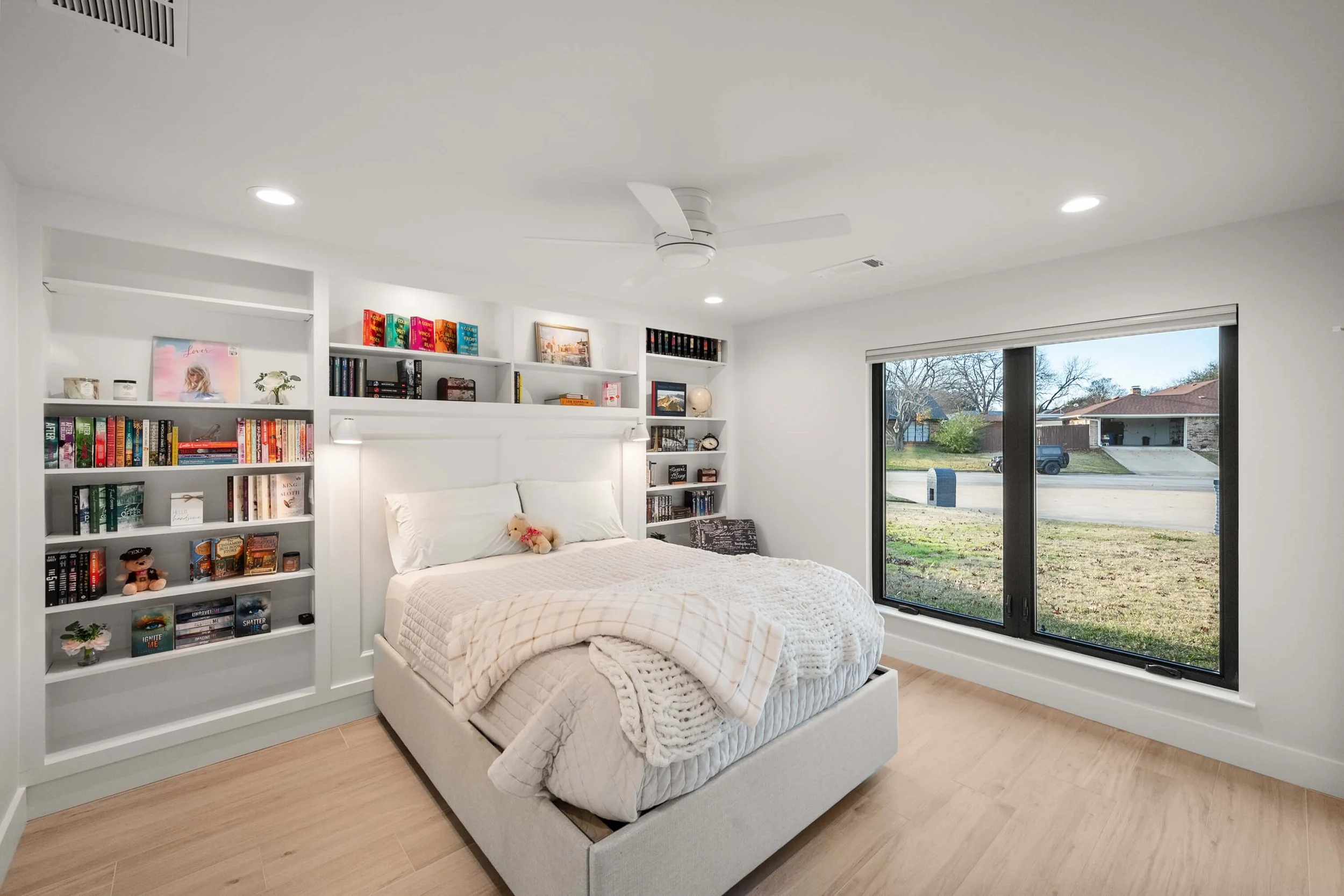 A bedroom with white walls, a large window, built-in shelves filled with books and decor, a white bed with pillows and a stuffed animal, and a ceiling fan.