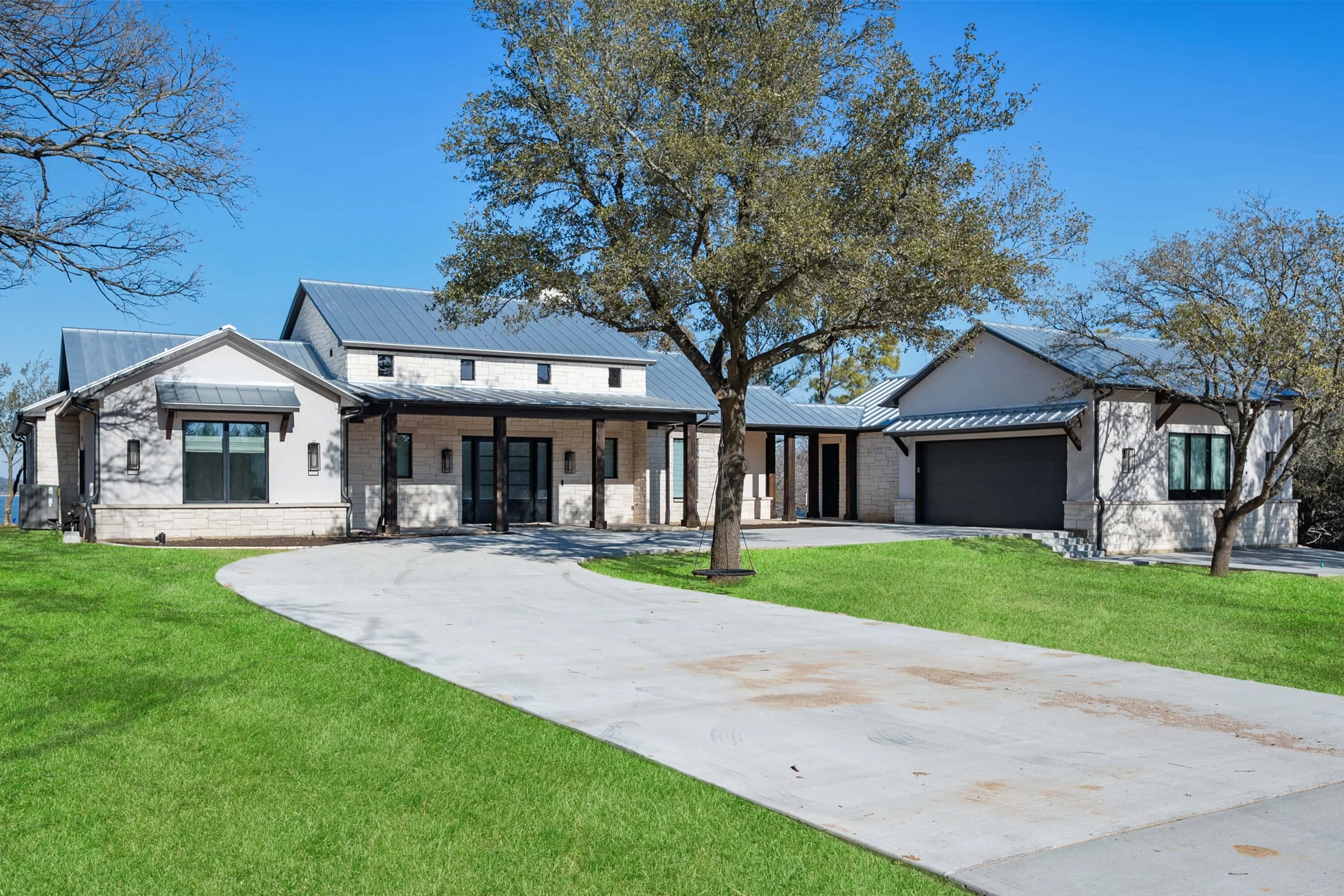 Modern house with metal roof, white brick exterior, black accents, surrounded by green lawn and trees, under a clear blue sky.