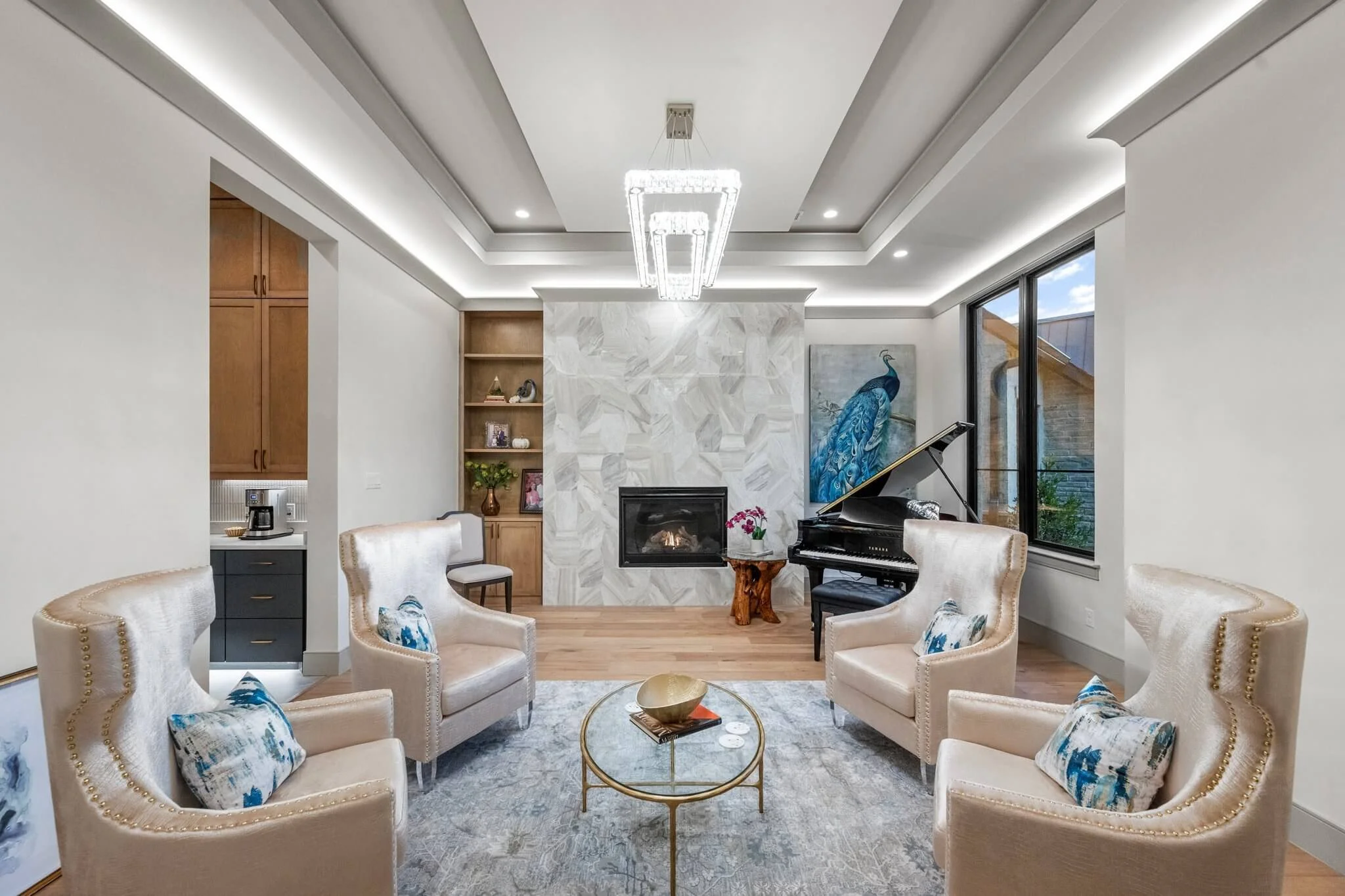 Living room with beige armchairs, a glass coffee table, white walls, wooden floor, fireplace, grand piano, peacock painting, and large windows in Colleville, TX.