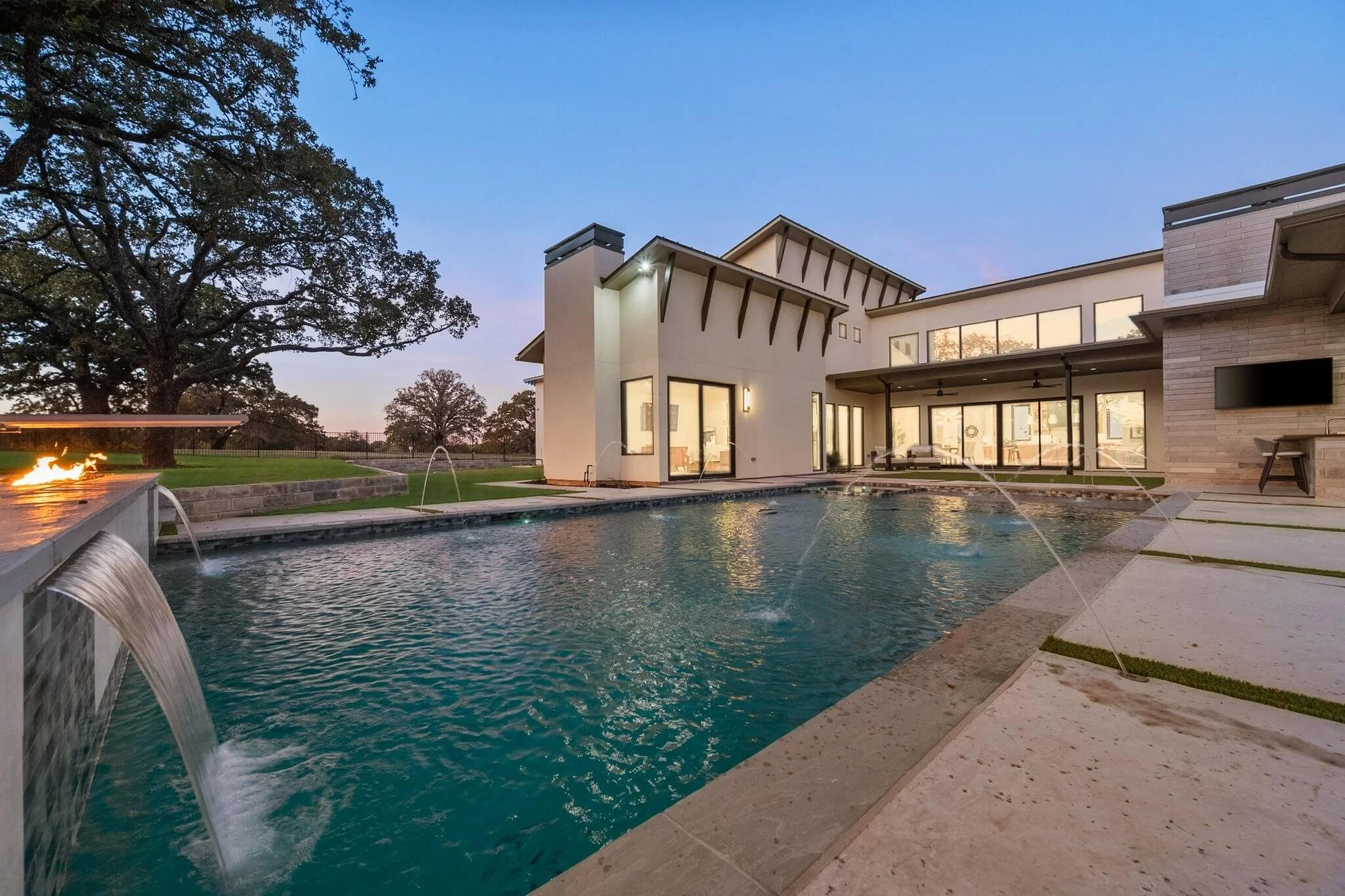 Modern house with large windows overlooking a swimming pool with water features, surrounded by a patio, trees, and an open yard at dusk.