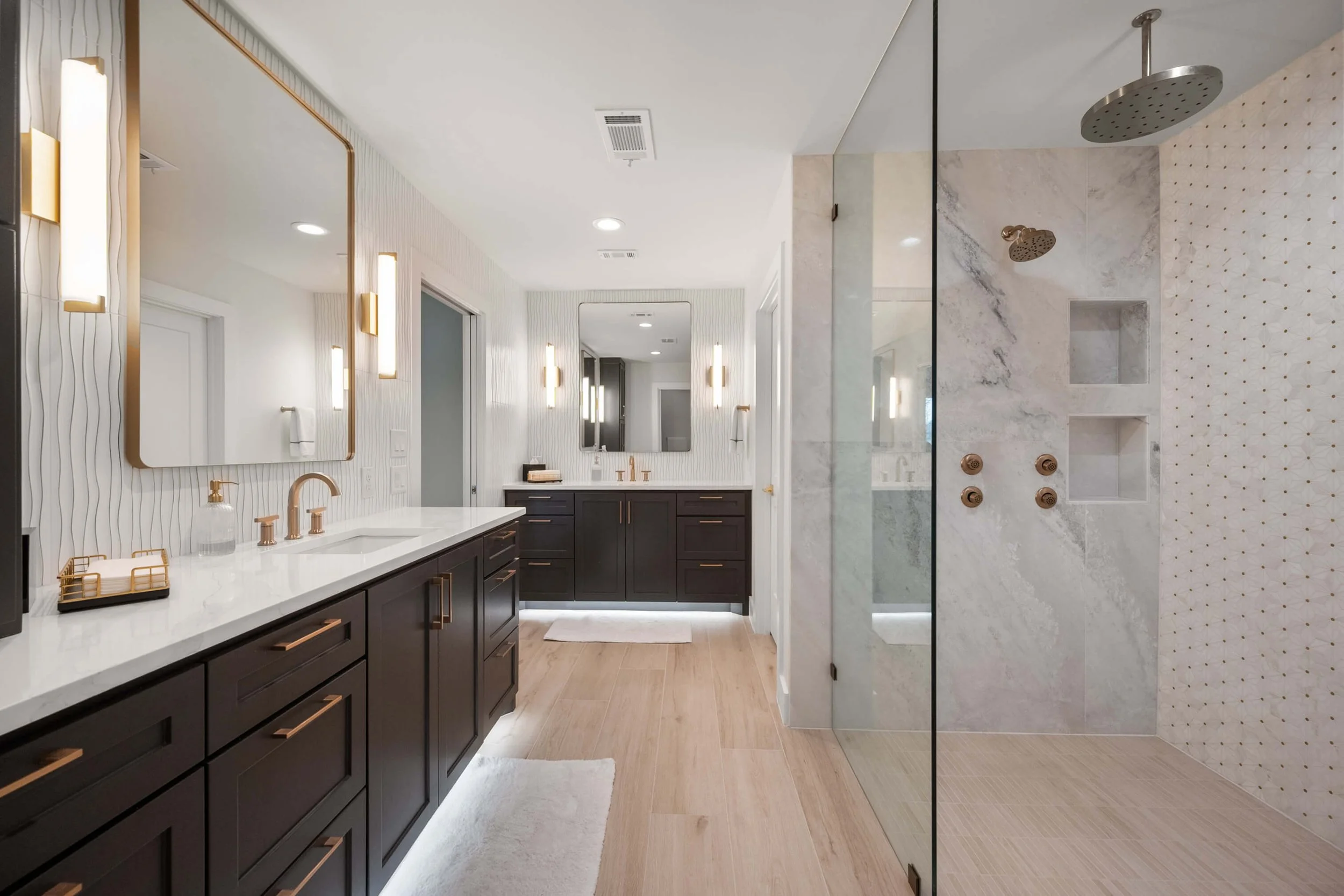 Modern bathroom with dual vanities, large mirrors, and black cabinetry; walk-in shower with marble walls, multiple showerheads, and built-in niches; wood flooring and minimal decor  built by Lakeland Custom Homes..