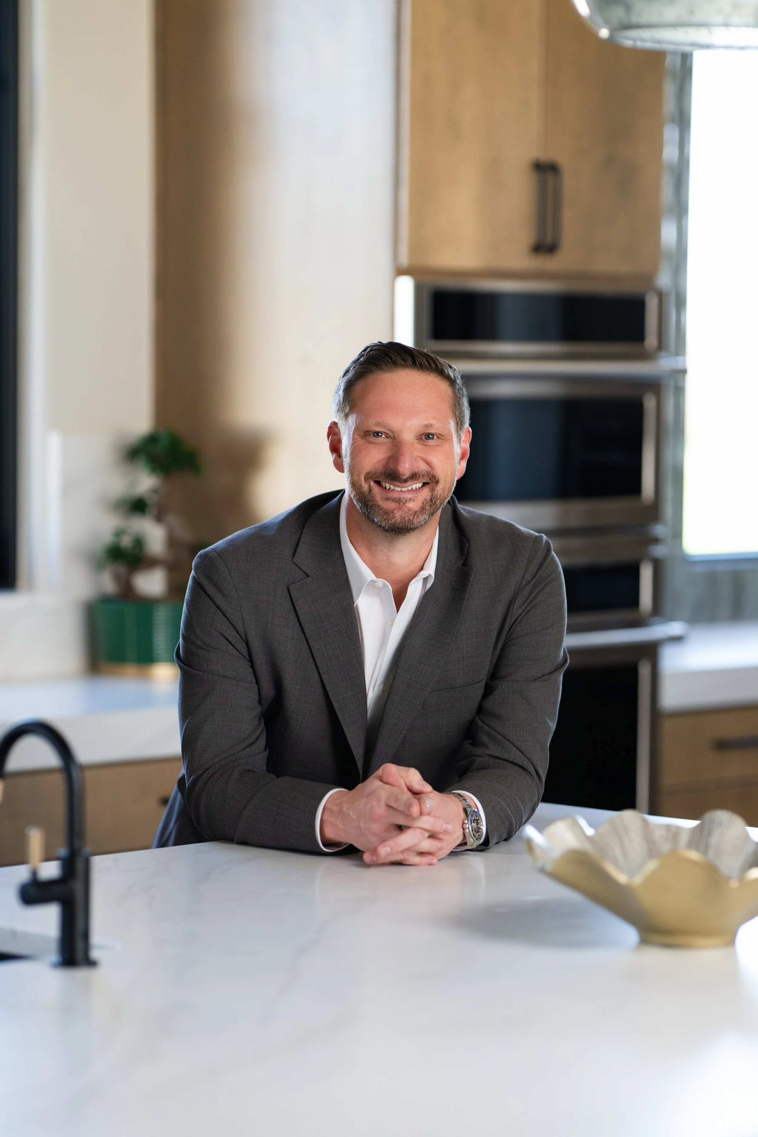 Nick Zuckerbrow, owner of Lakeland Custom Homes, in a gray suit sitting at a kitchen counter, smiling with hands clasped.