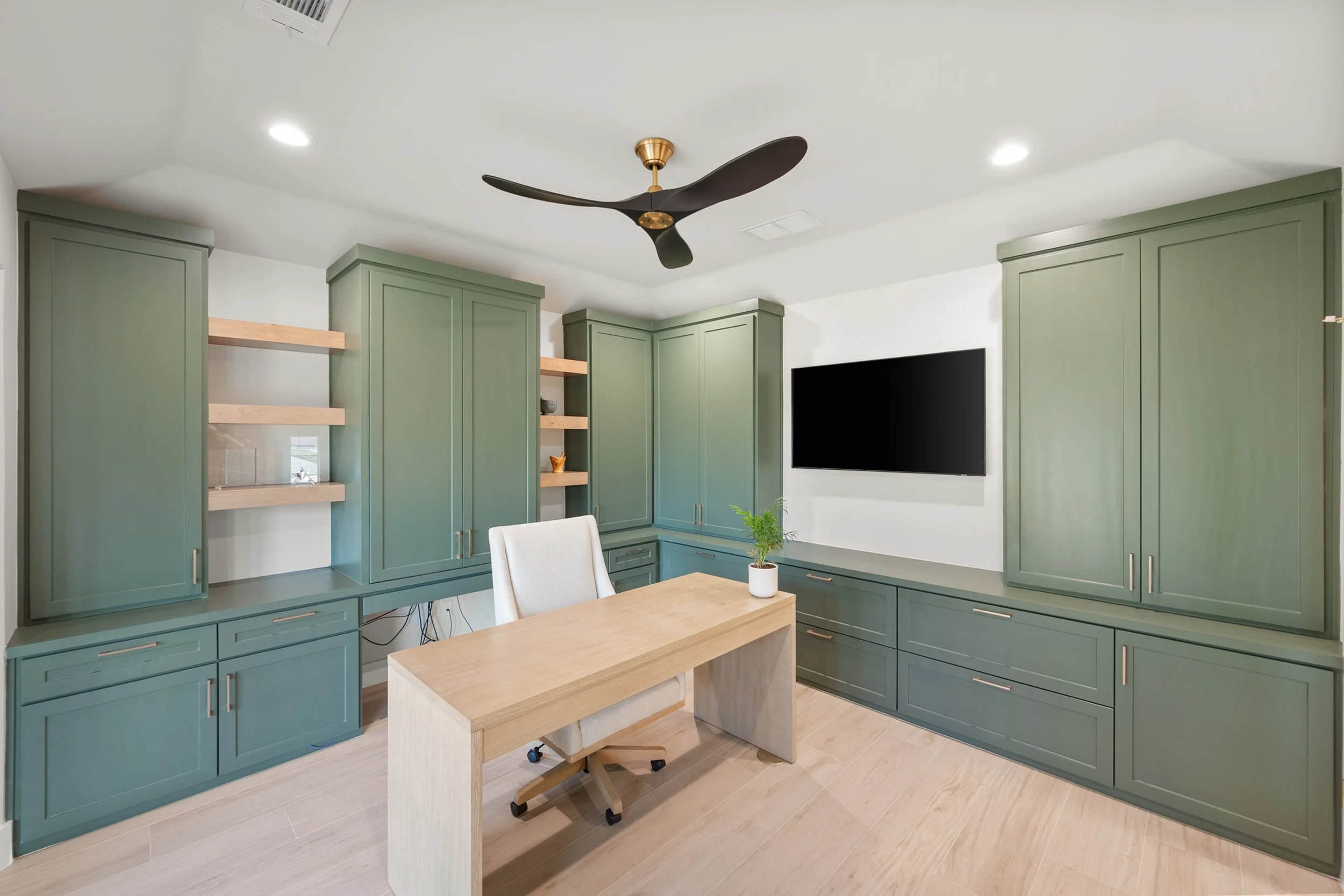Modern home office with green cabinetry, a wooden desk, a beige desk chair, a black ceiling fan, a potted plant, and a wall-mounted television.