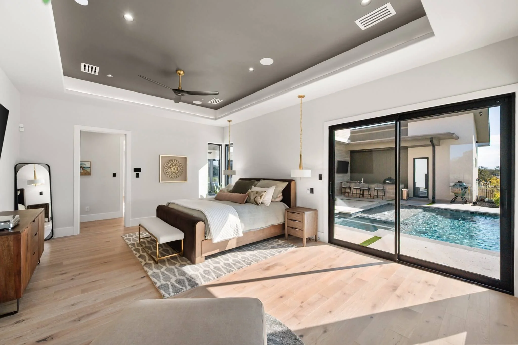 Modern bedroom with large sliding glass door leading to a pool area outside. The room has light wood flooring, a bed with neutral bedding, hanging pendant lights, and a ceiling fan. The decor is minimalist with white walls and simple artwork.