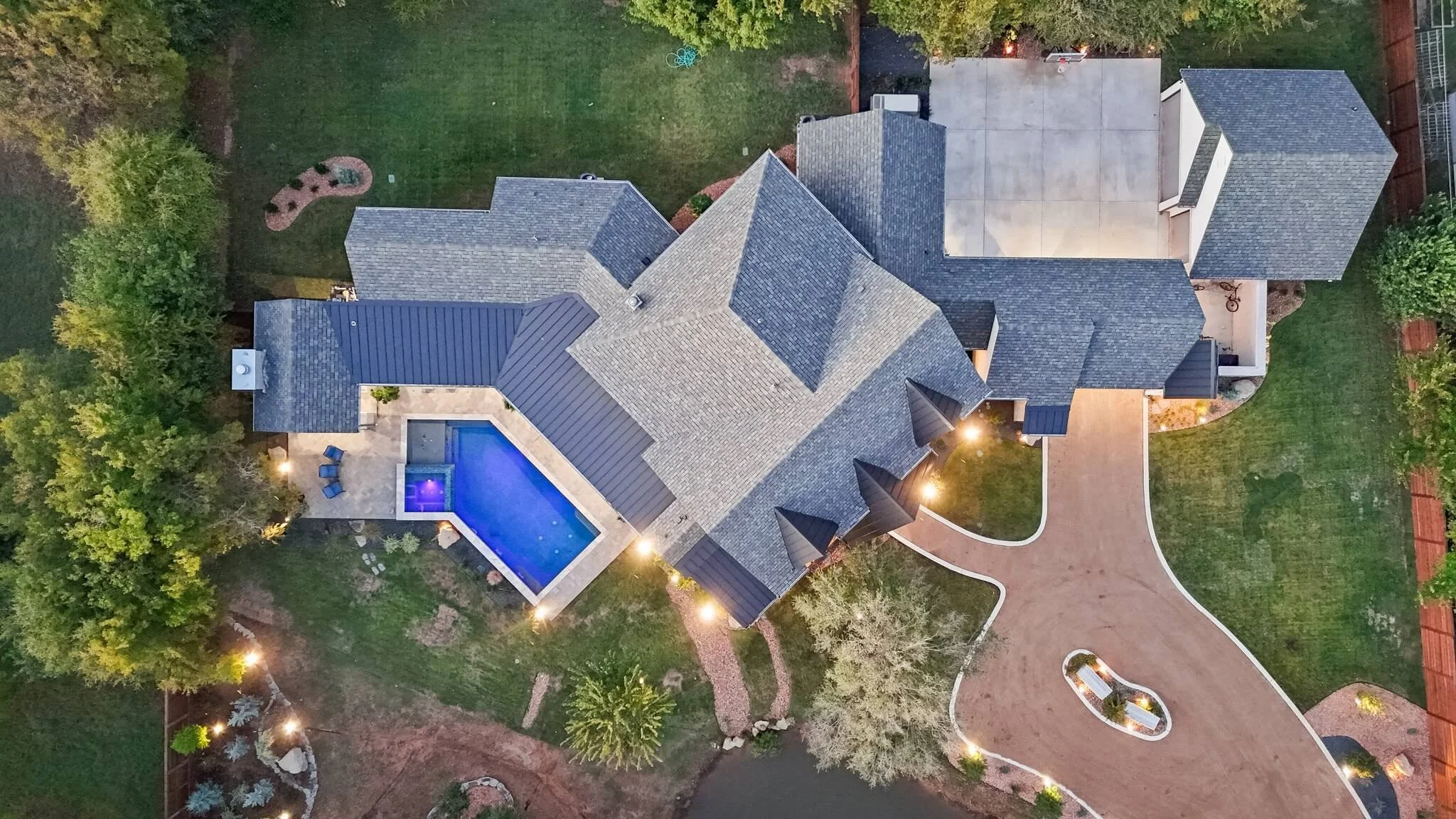 An aerial view of a residential house with a backyard, swimming pool, garden, and paved driveway illuminated at dusk.