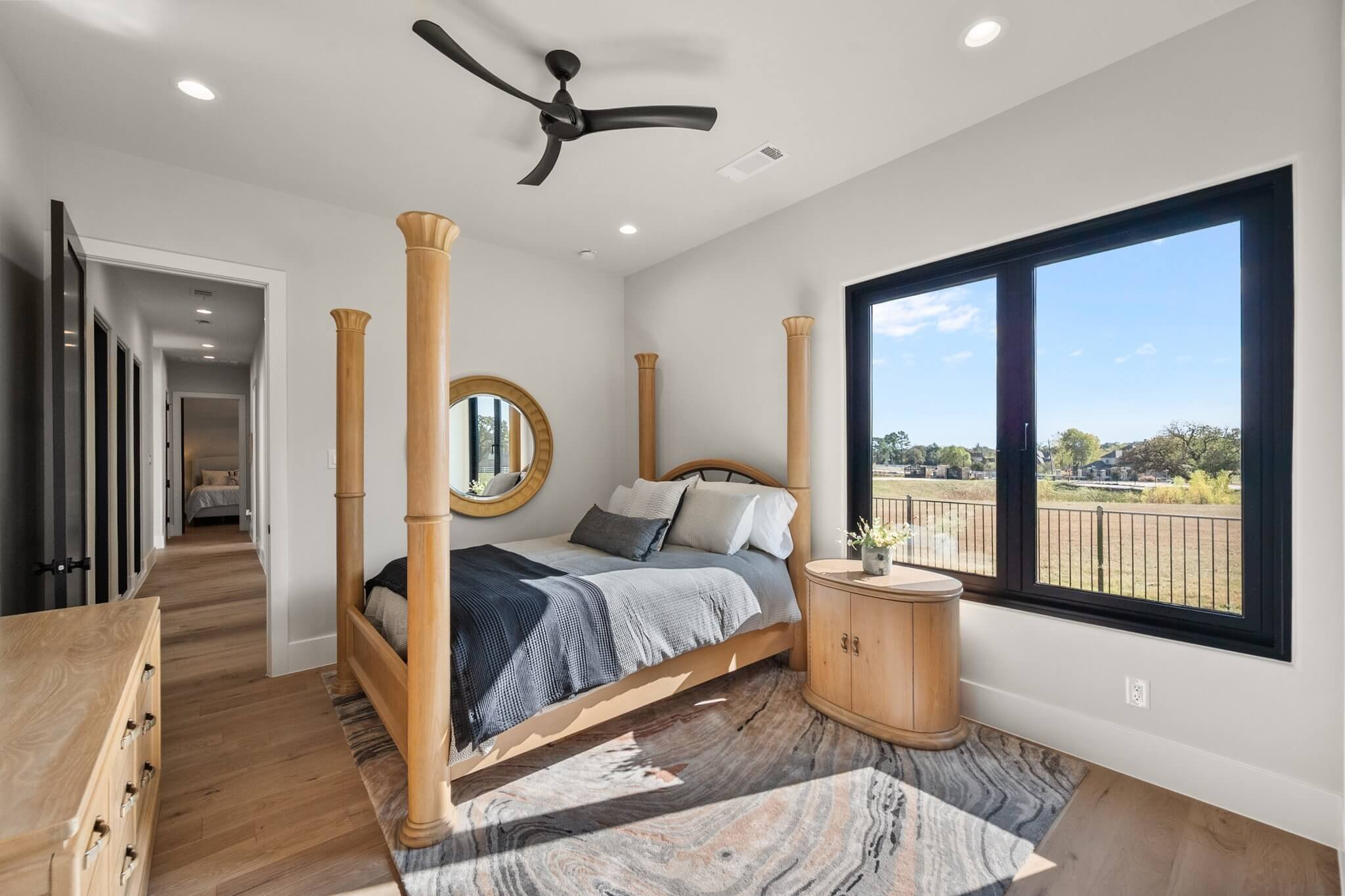 A bedroom with a four-poster wooden bed, gray bedding, and white pillows, next to a window with a view of a field and trees, a small wooden nightstand with a potted plant, a mirror on the wall, and a hallway leading to another room.