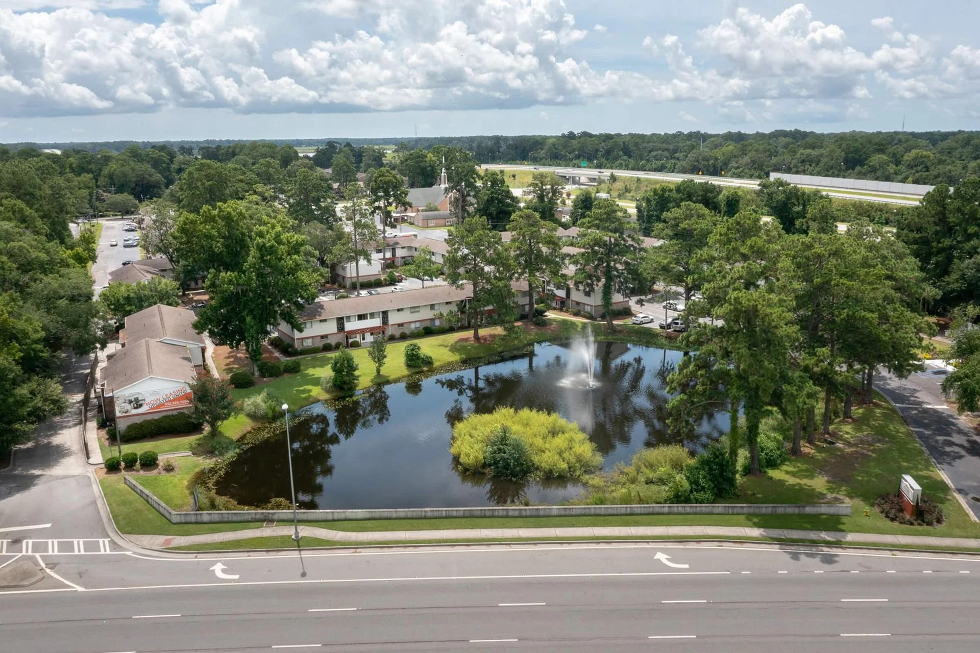An aerial view of a pond with a fountain, surrounded by trees and residential buildings, with a highway in the distance under a cloudy sky.