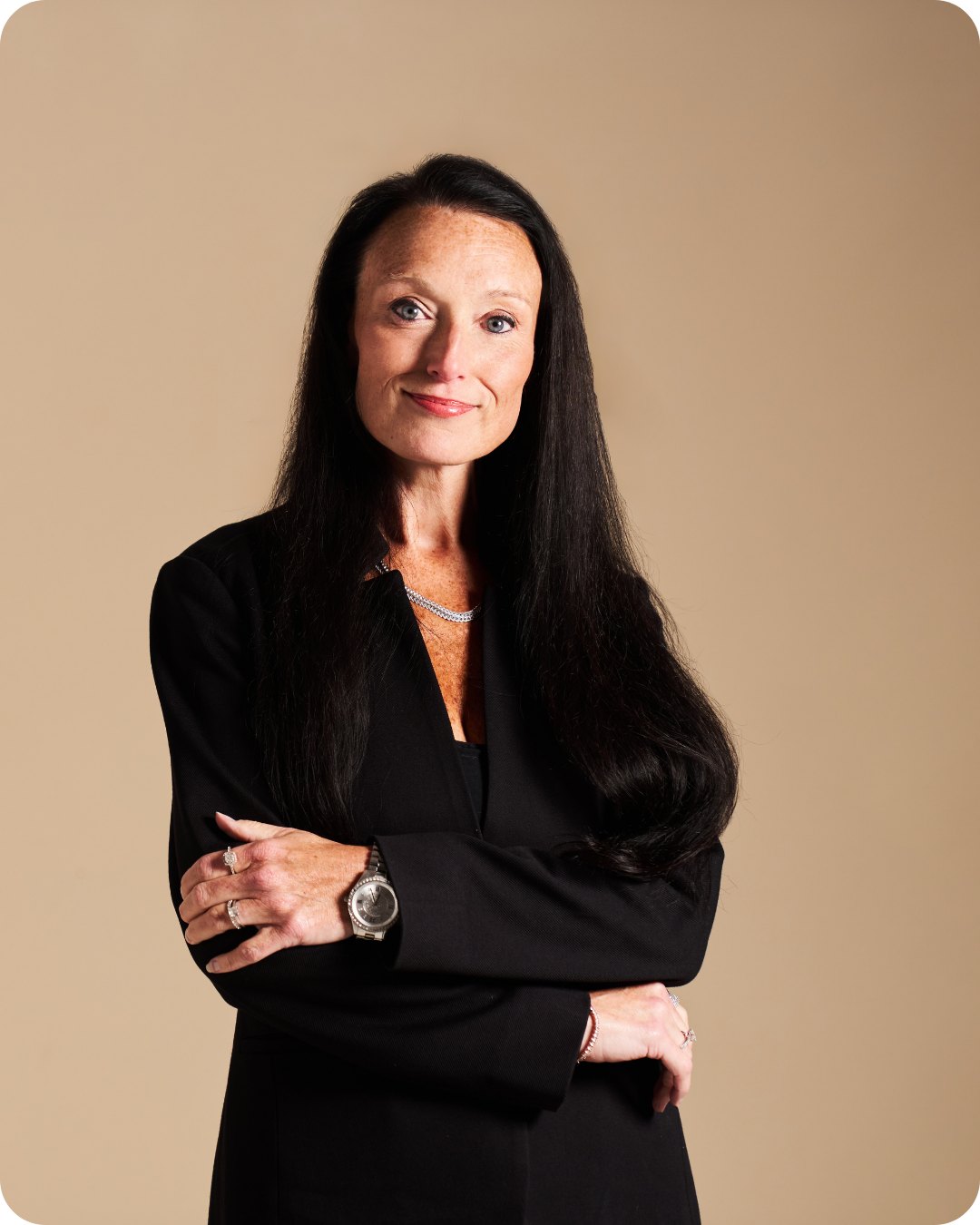 A professional woman with long dark hair, wearing a black blazer, jewelry, and a watch, standing with arms crossed against a beige background.