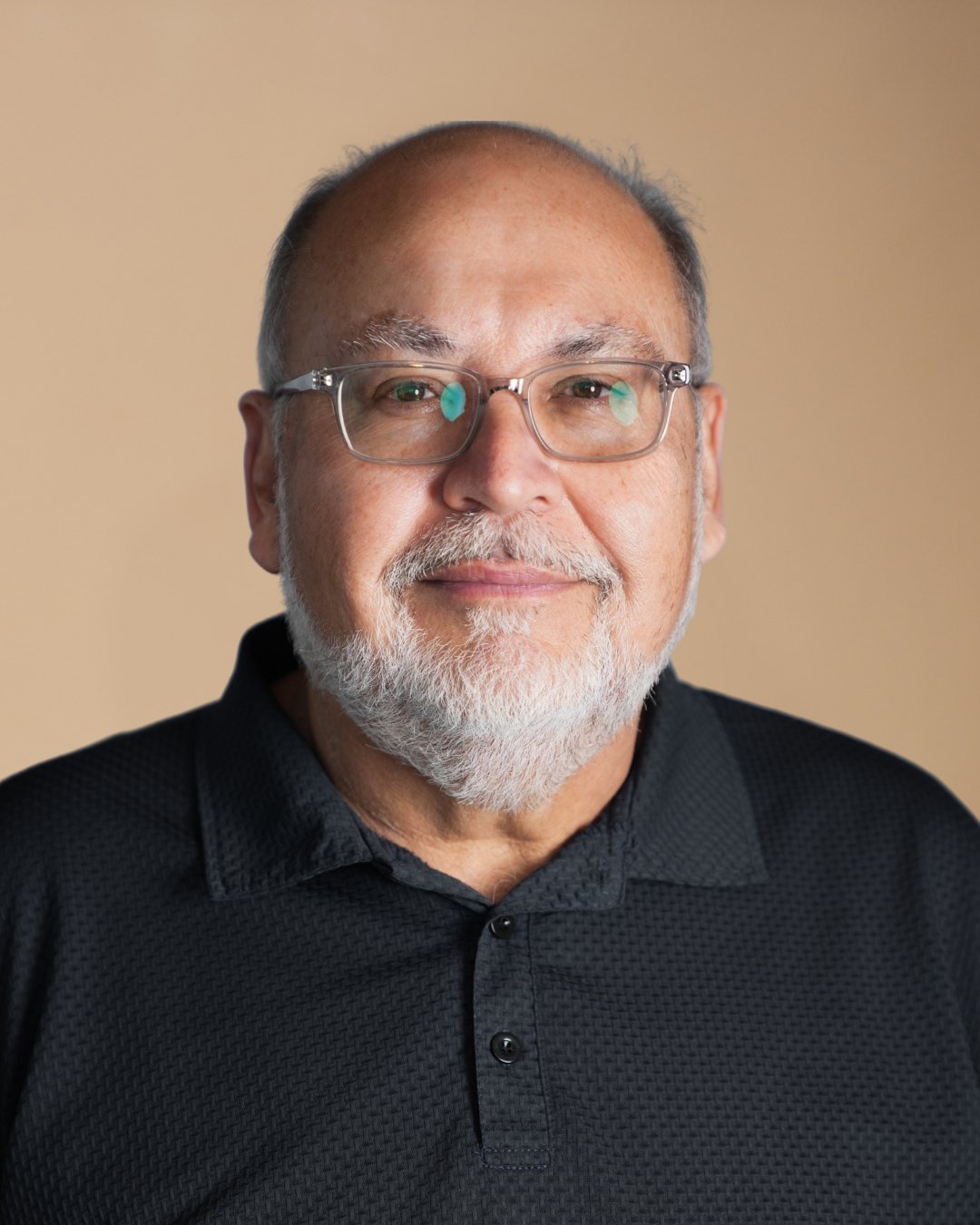 A middle-aged man with glasses, a beard, and short gray hair, wearing a black collared shirt, smiling against a beige background.