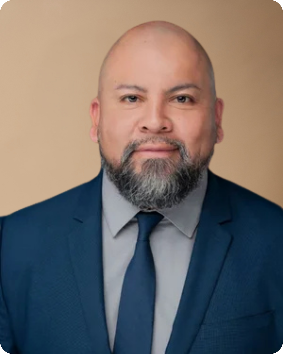 A man with a beard and mustache wearing a dark blue suit, light gray shirt, and dark blue tie, against a neutral background.