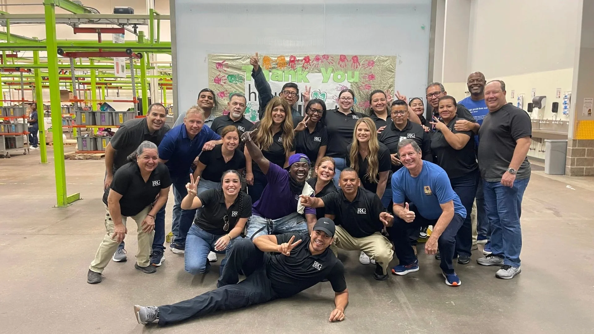 A group of diverse people smiling and posing together in a warehouse or factory with a decorated 'Thank You' banner in the background, some making peace signs and other gestures of celebration.