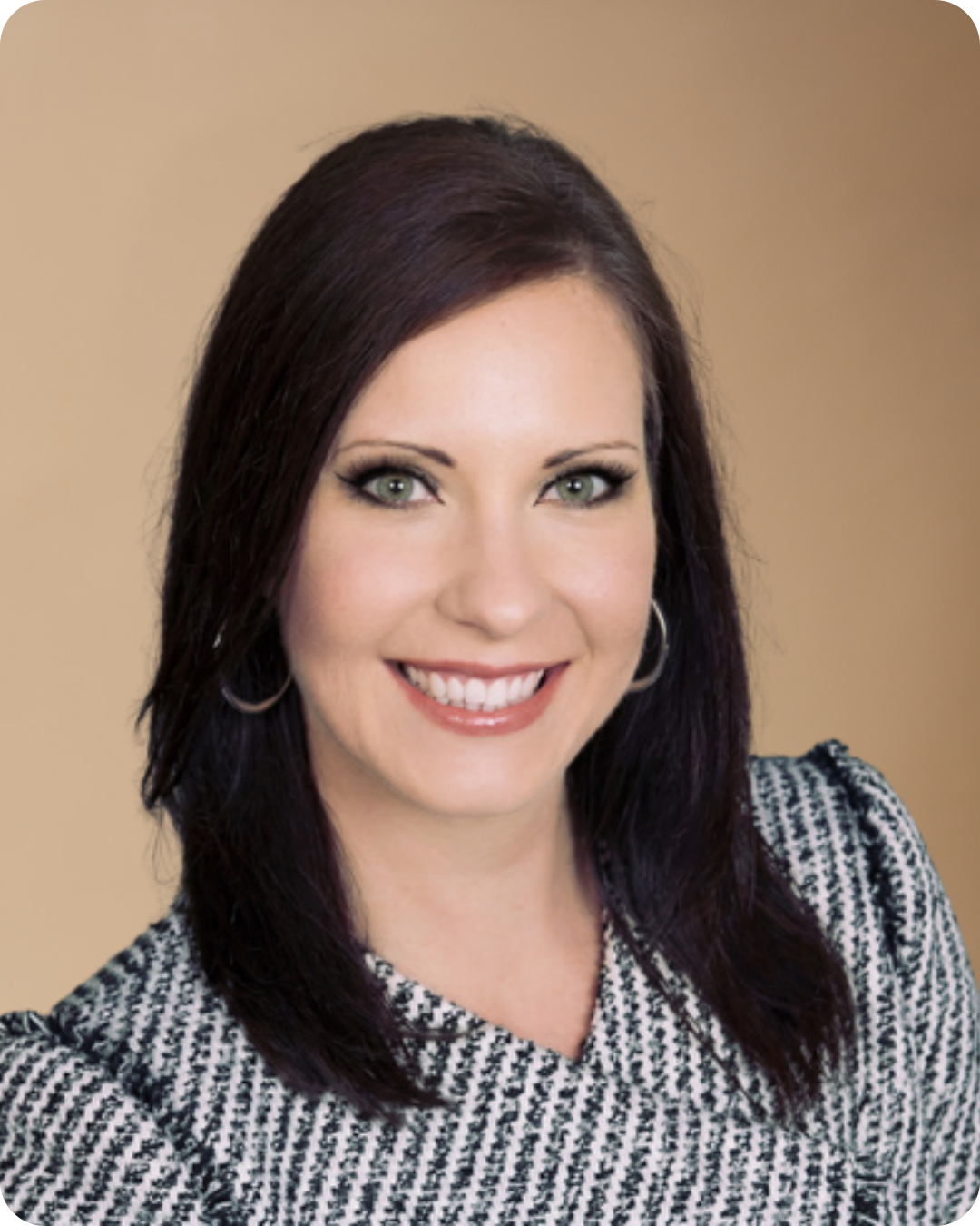 A smiling woman with shoulder-length dark hair, green eyes, and hoop earrings, wearing a black and white patterned top, against a beige background.