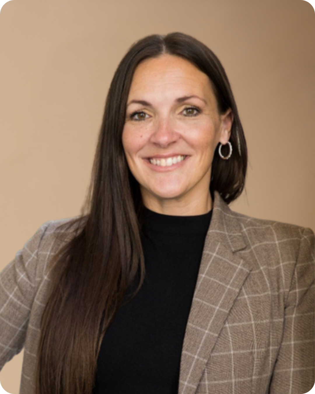 A professionally dressed woman with long dark hair, smiling, wearing a brown plaid blazer and hoop earrings, standing against a beige background.